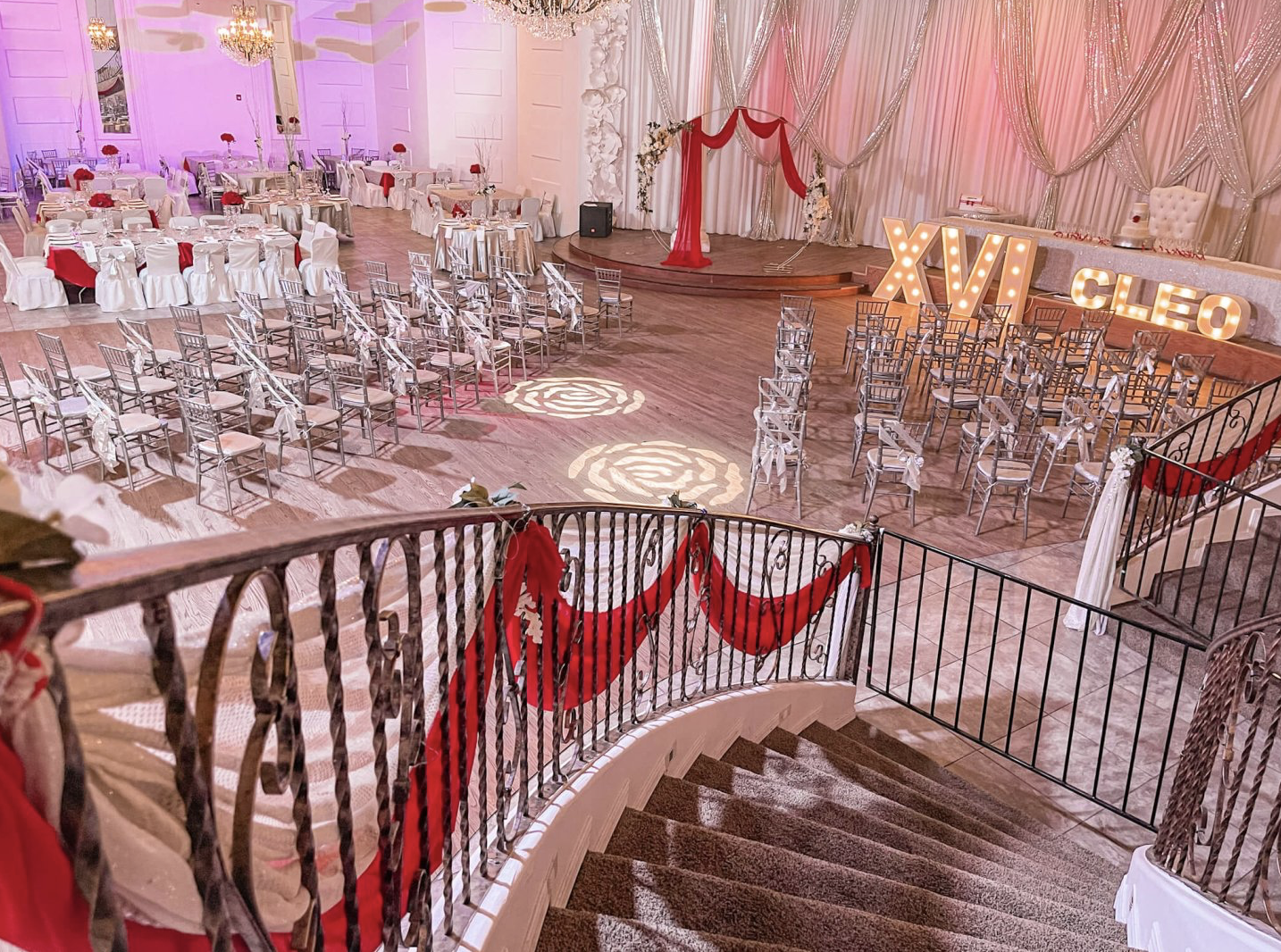 Quinceañera celebration setup featuring white tables and chairs, red draping, and a decorated staircase.