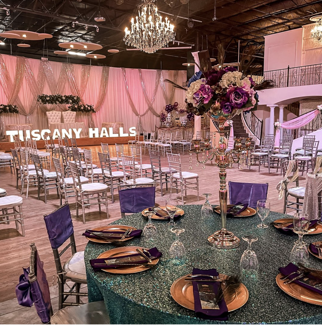 Colorful event setup featuring decorated tables, floral centerpieces, clear chairs, and pink and purple lighting.