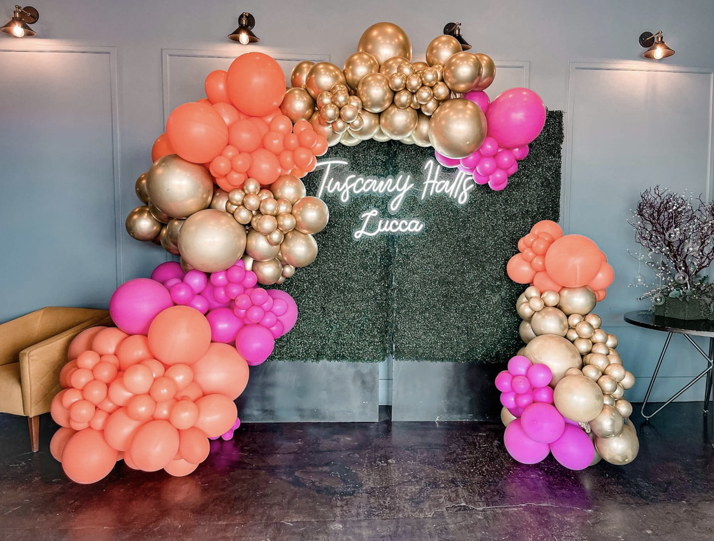 Balloon display featuring pink, peach, white, and metallic balloons surrounding a greenery backdrop.
