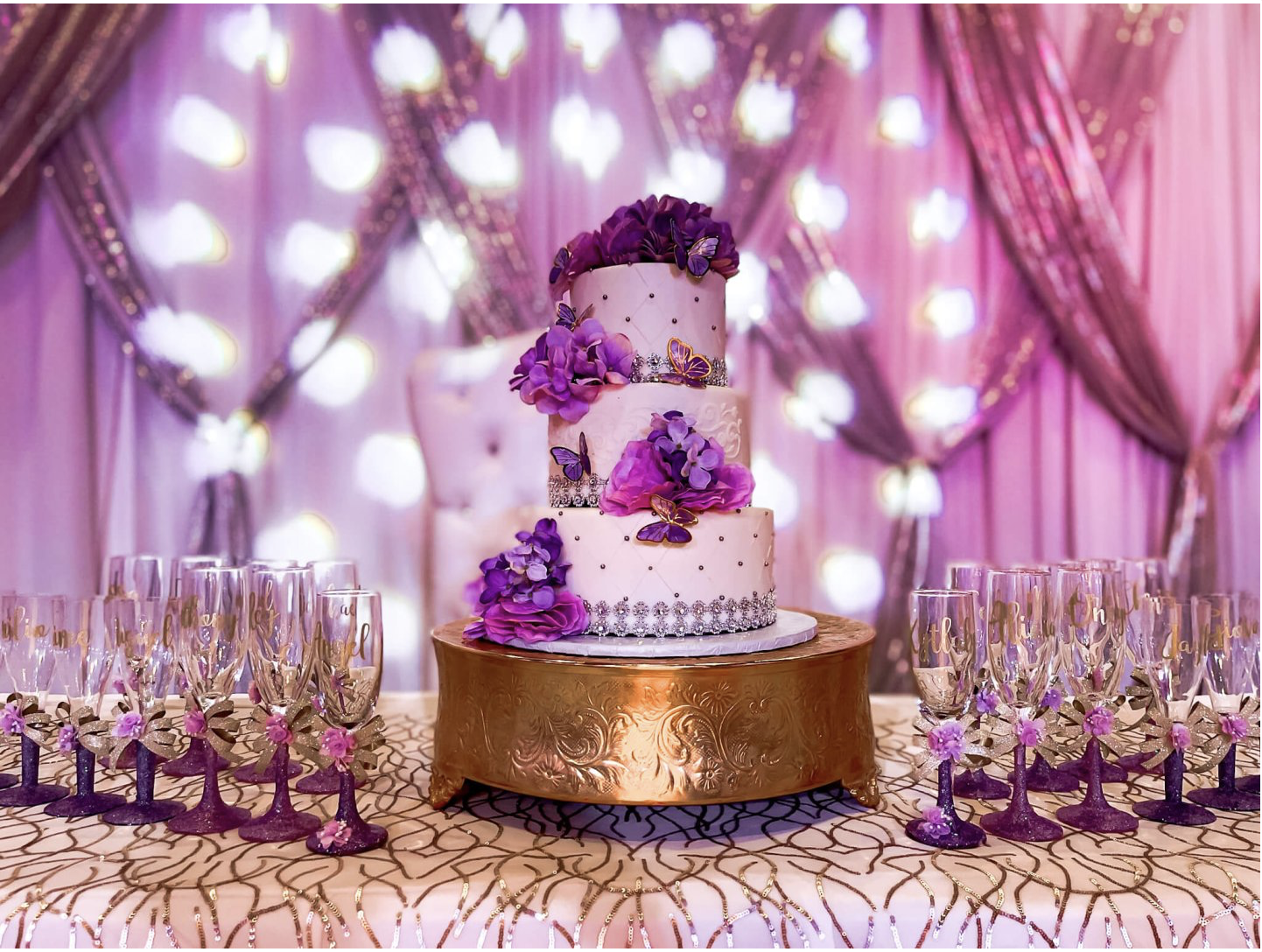 Decorated cake display featuring a purple cake, draped backdrop, soft lighting, and surrounding desserts.