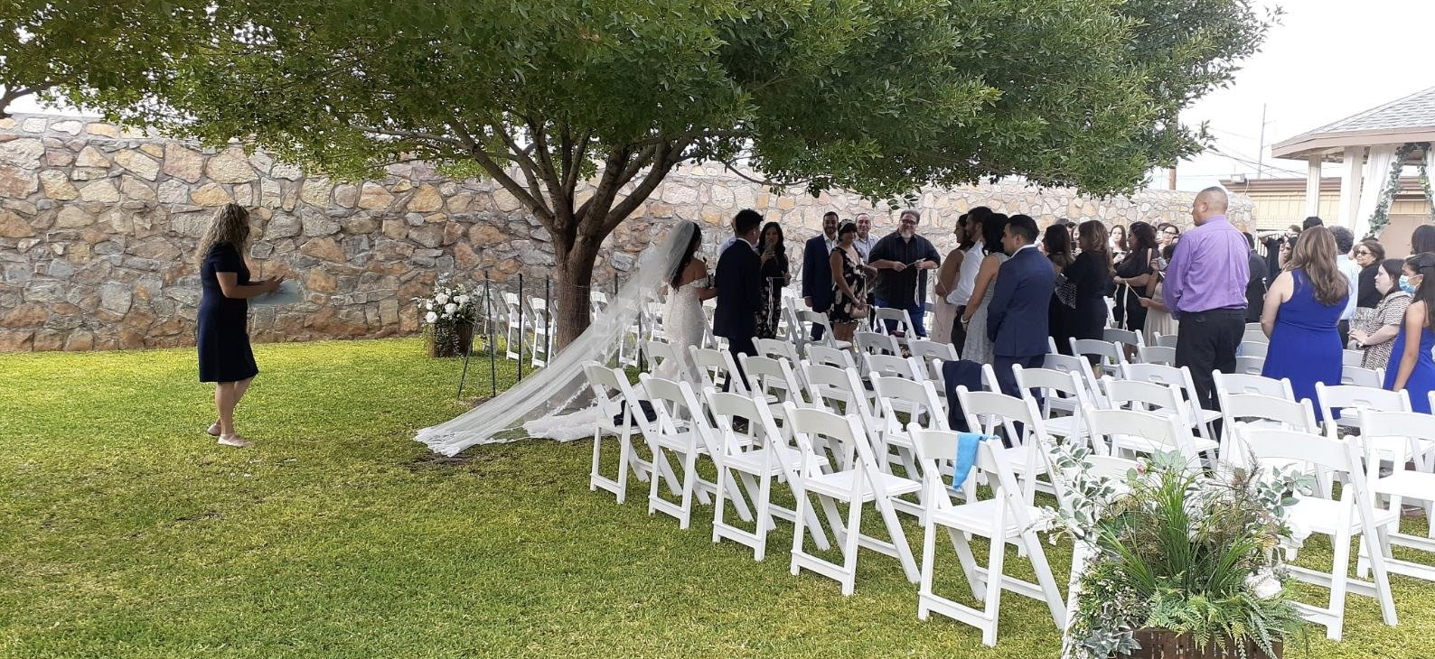 Outdoor ceremony setup featuring rows of white chairs on the lawn beneath a large shade tree.