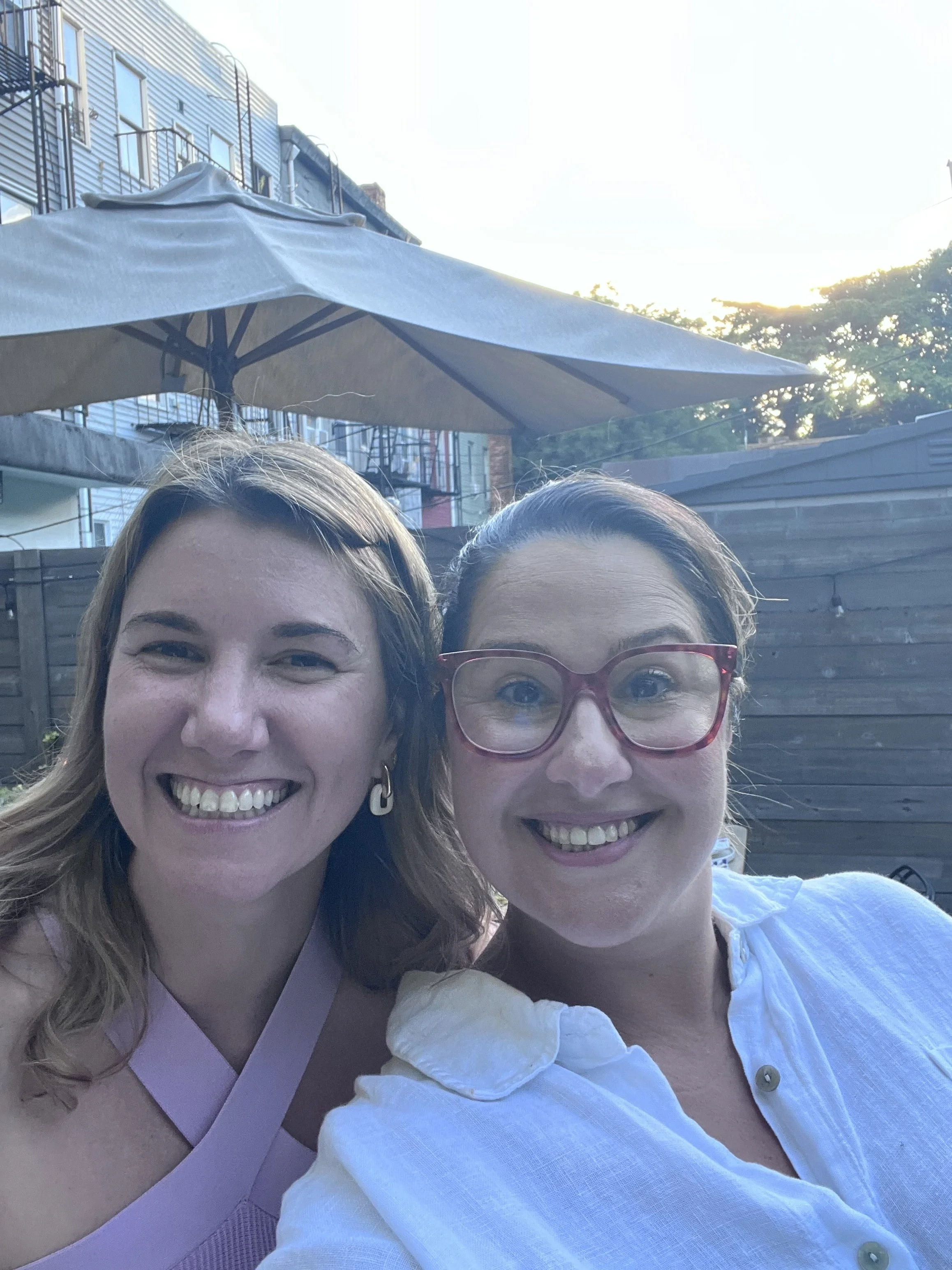 Two women smiling outdoors on a sunny day, with an umbrella, in a backyard setting with apartment building and trees in the background.