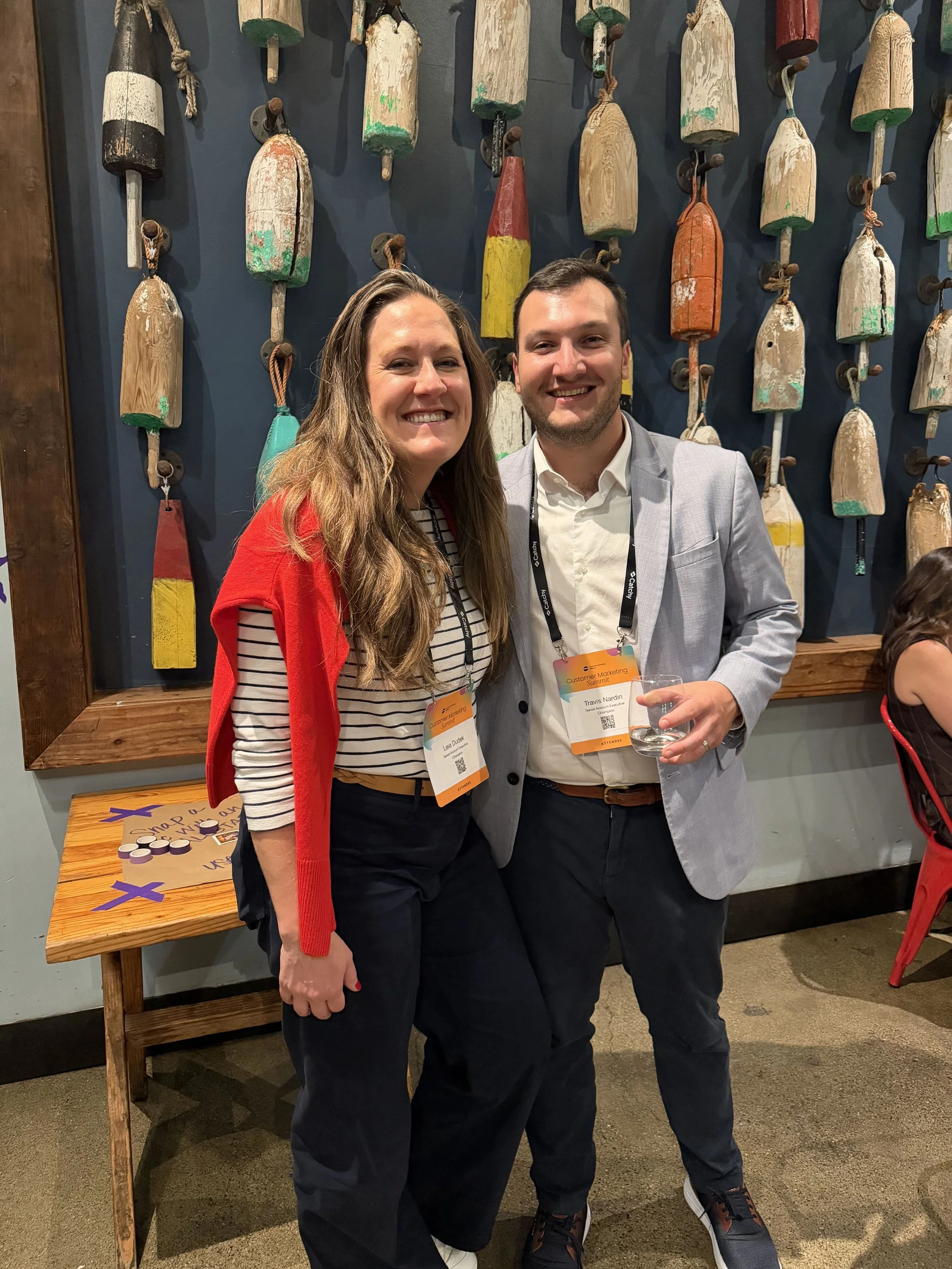 A woman and a man smiling and posing for a photo at a conference, standing close together. The woman is wearing a striped shirt with a red jacket, and the man is dressed in a light gray blazer and dark pants. Both have conference badges hanging around their necks.