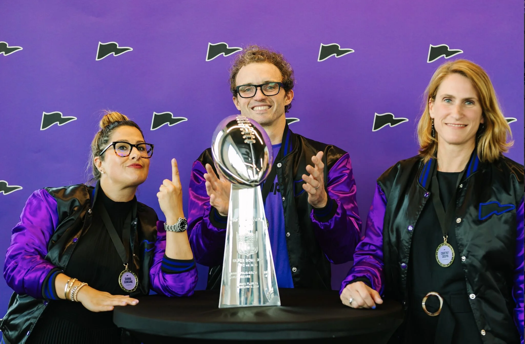 Three people standing behind a table with a trophy, wearing black jackets with purple accents, smiling, with a purple background featuring black and white logos.