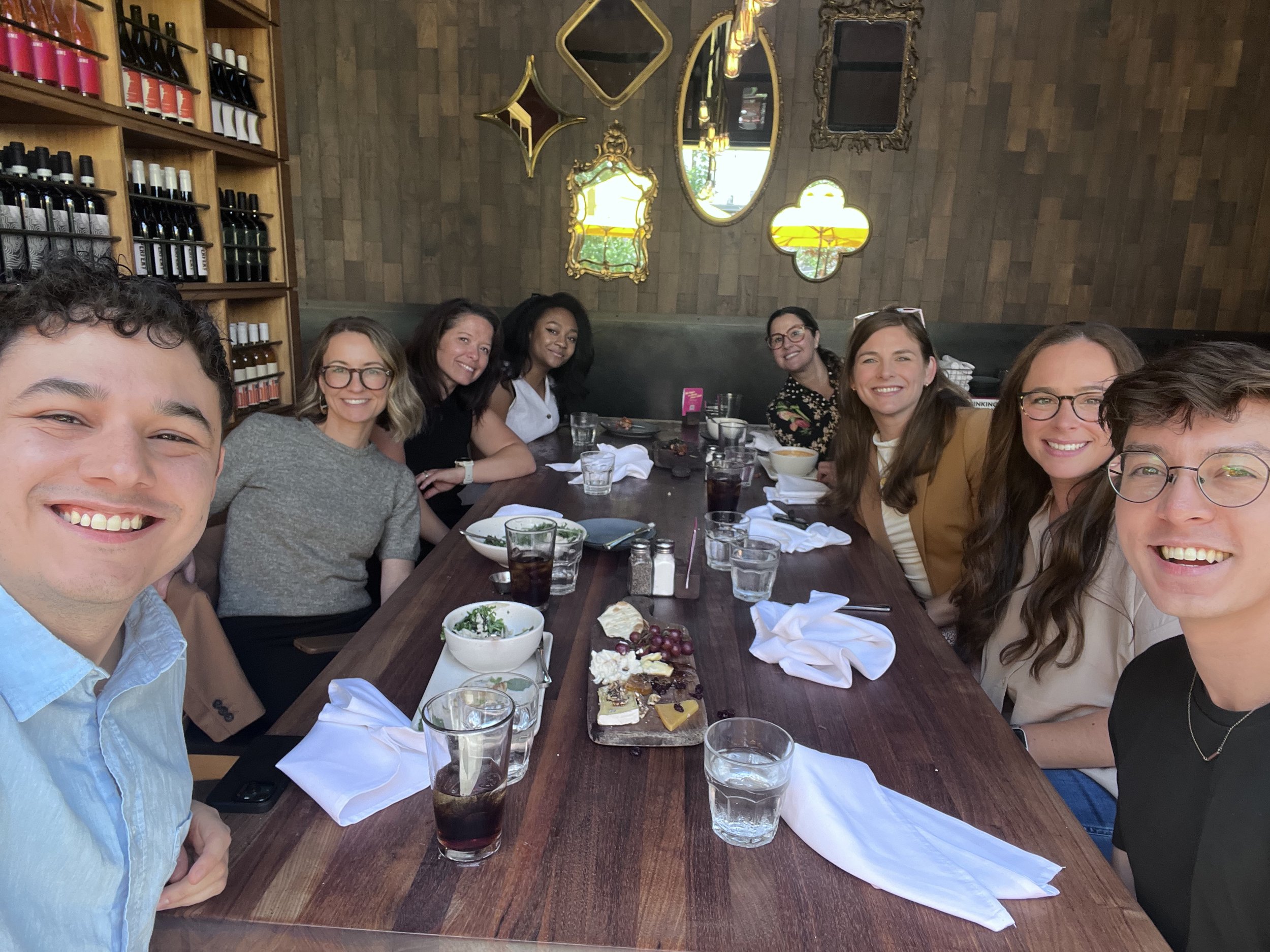 A group of eight friends sitting around a dining table at a restaurant, smiling at the camera. The table has various plates of food, drinks, and utensils, with a wooden wall decorated with mirrors and wine bottles in the background.