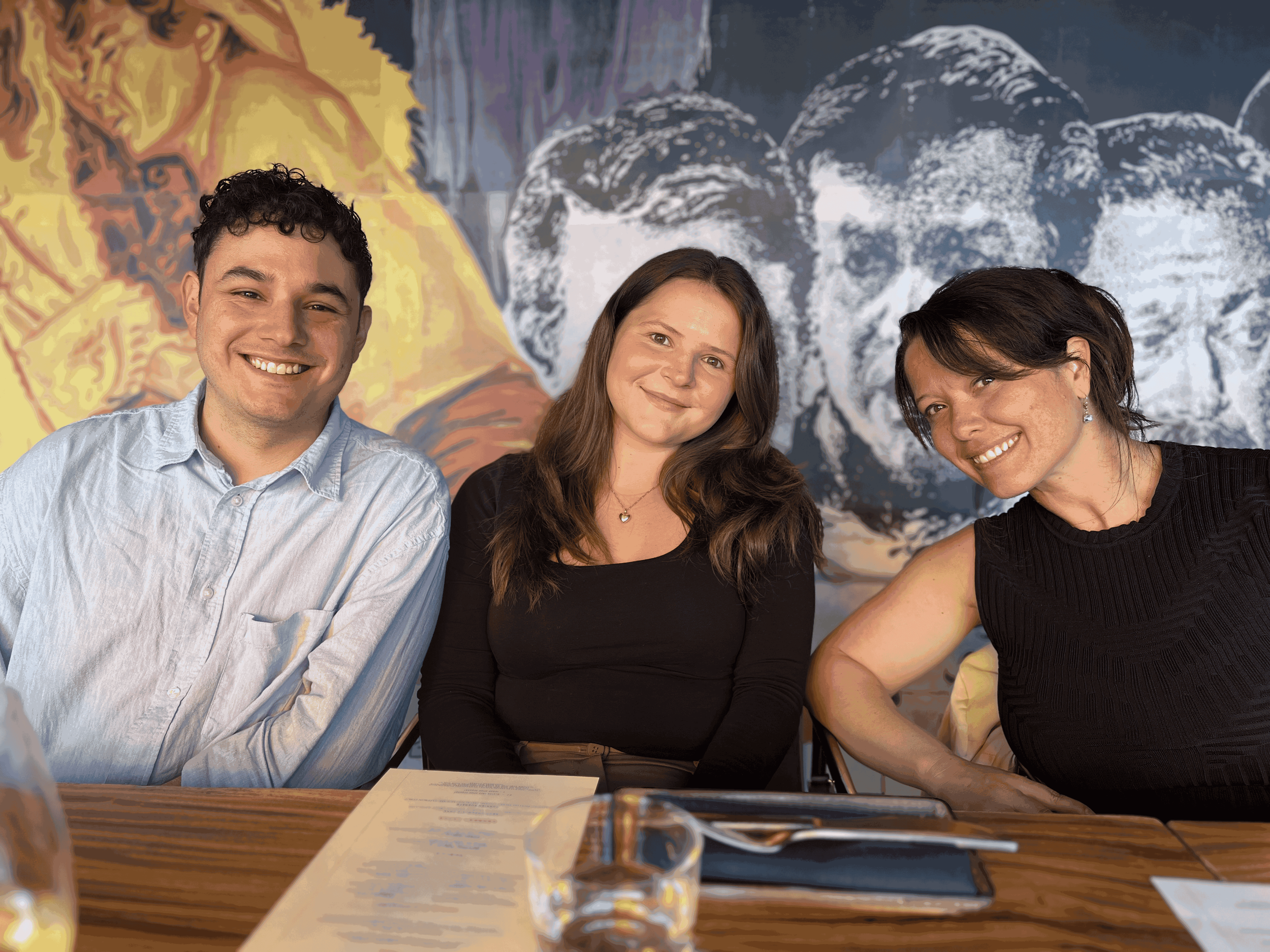 Three people sitting at a table with a document, glasses, and writing tools, in front of a mural featuring artistic faces. They are smiling and looking at the camera.