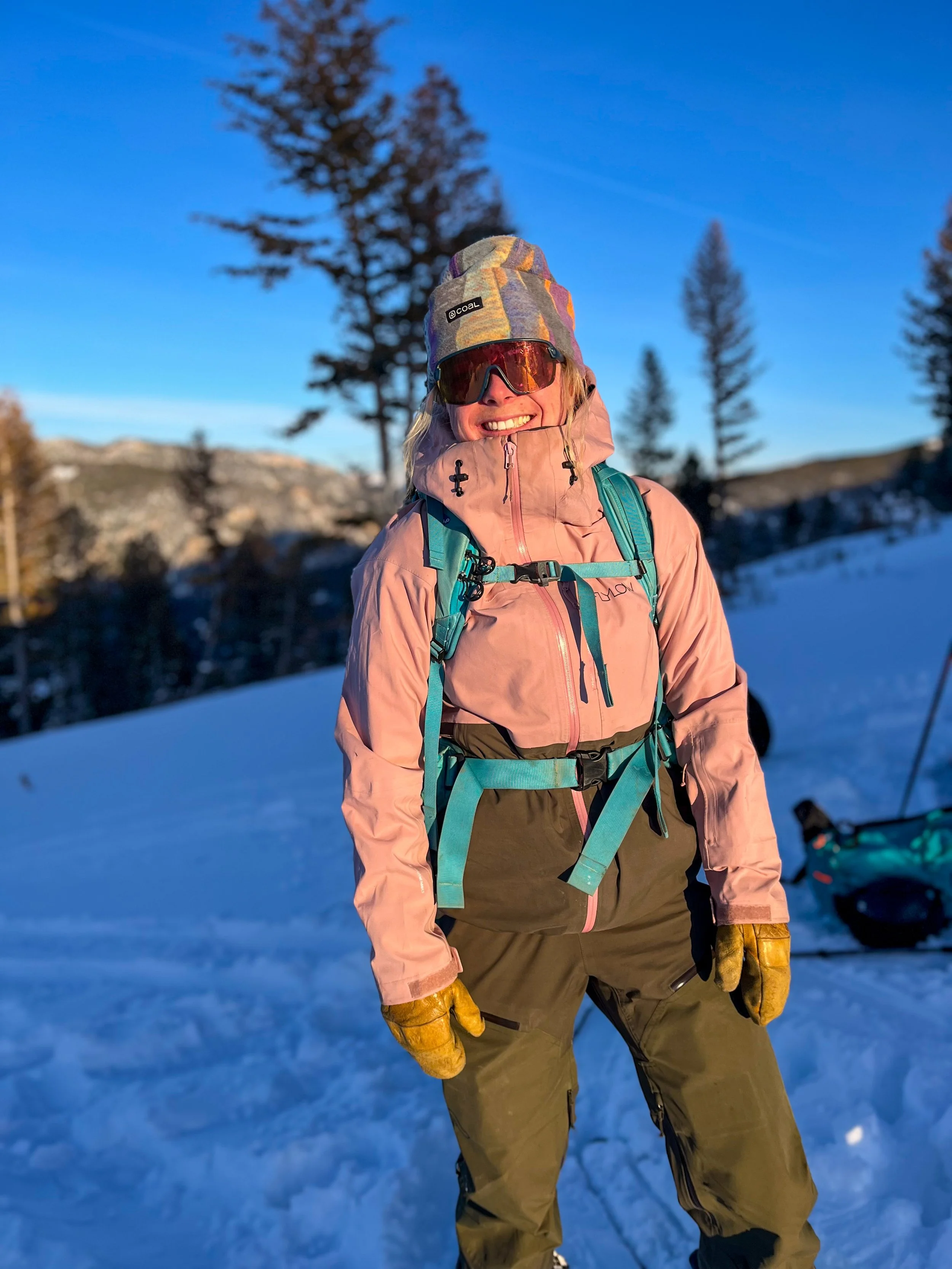 A woman dressed in winter gear, including a colorful beanie, reflective sunglasses, a pink jacket, and a backpack, standing on a snowy landscape with tall pine trees and a blue sky in the background.