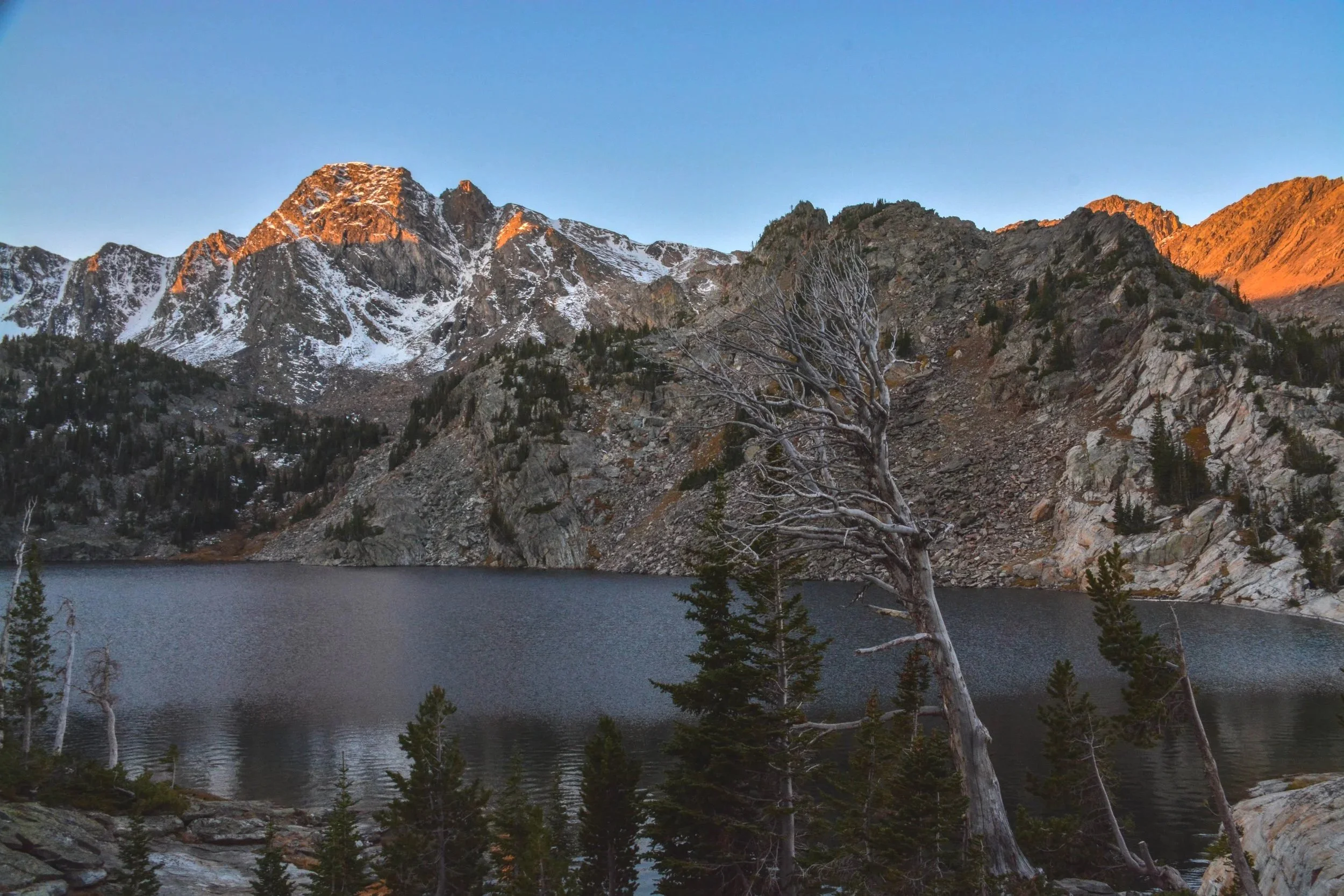 Mountain landscape with snow-capped peaks, a calm lake in the foreground, and a clear blue sky at sunset.