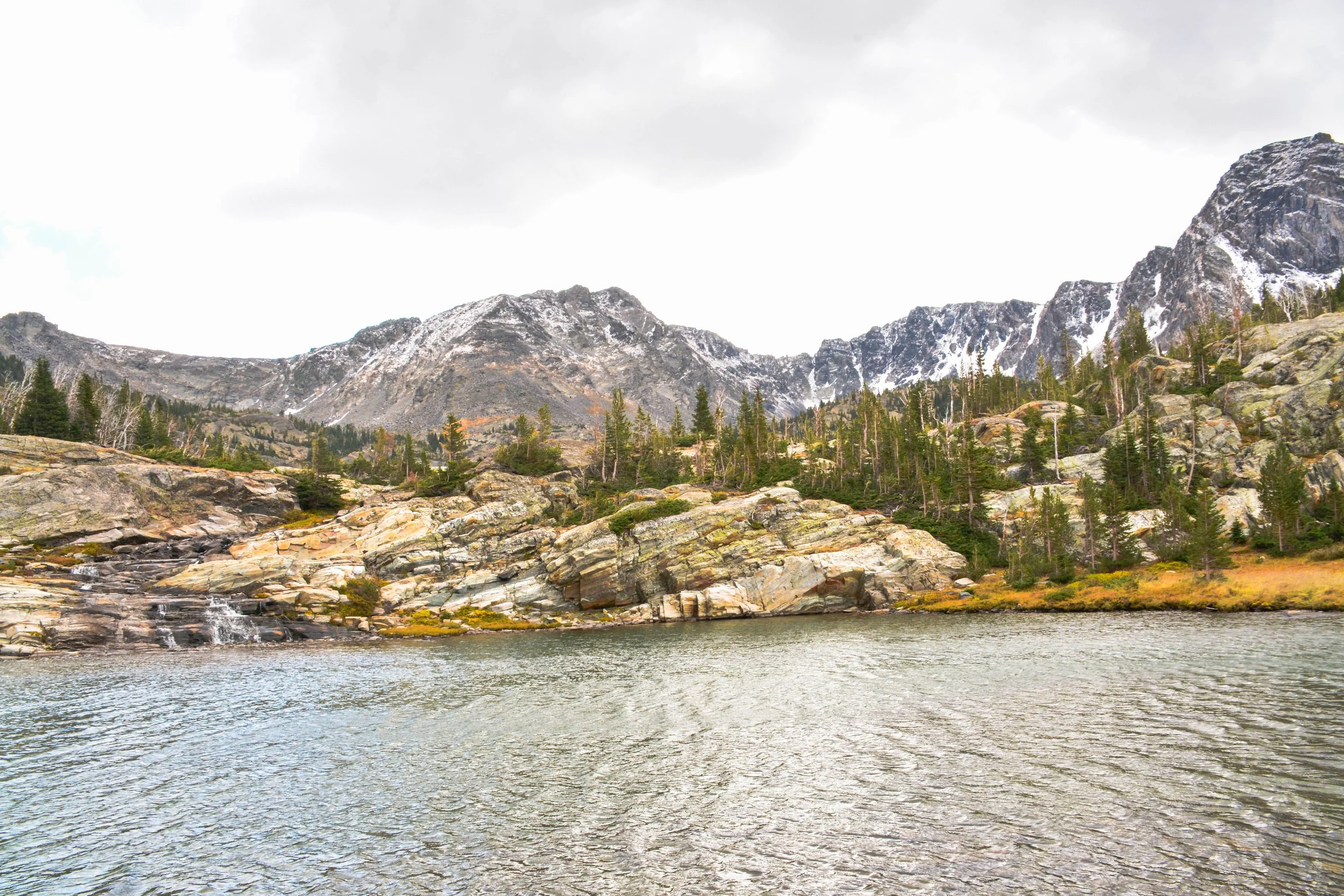Mountain landscape with rocky slopes, evergreen trees, and a body of water in the foreground under cloudy sky.