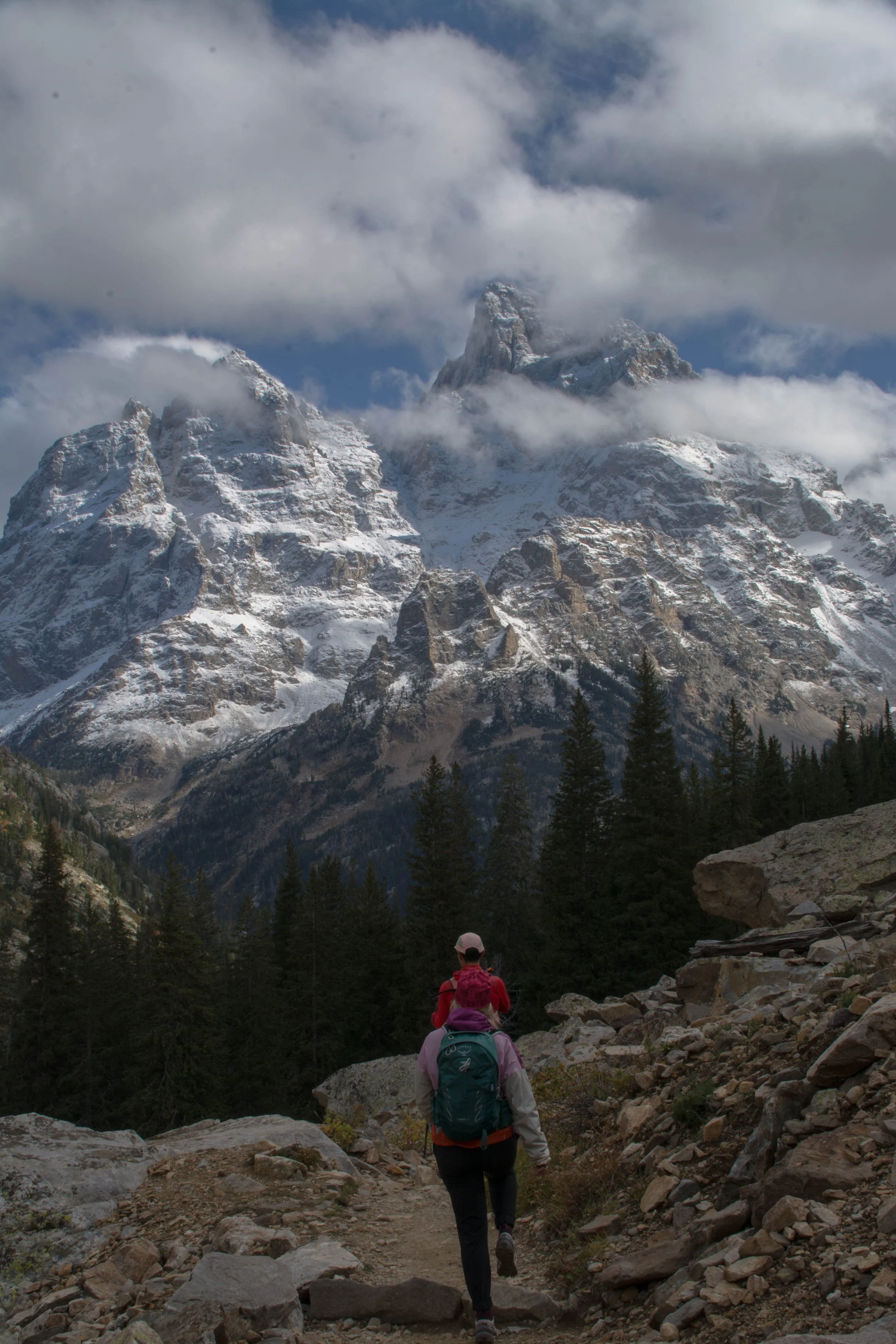 Hiker with backpack walking on rocky trail in forested mountain landscape with snow-capped peaks and cloudy sky in the background.
