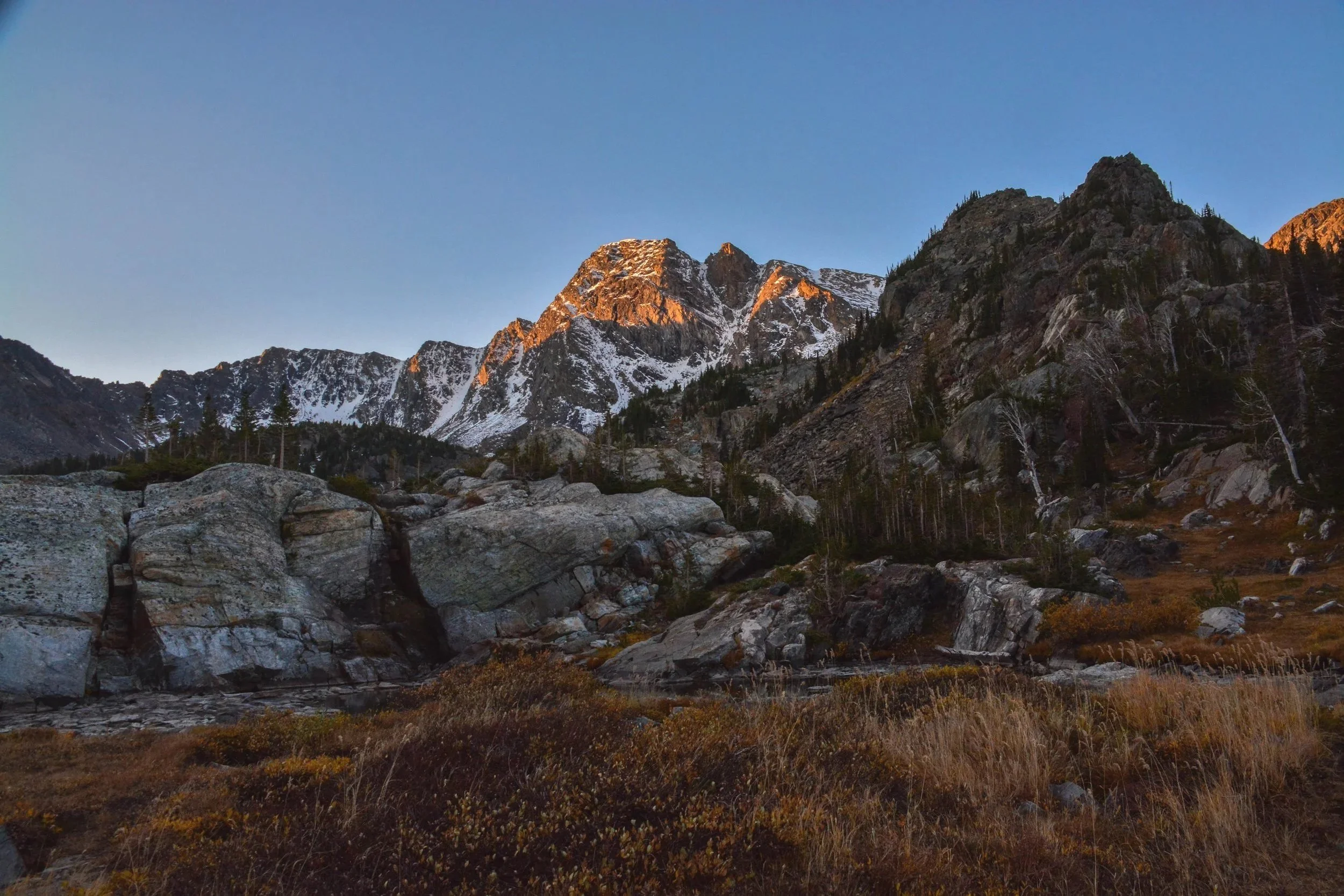 Mountain landscape at sunset with snow-capped peaks, rocky foreground, and sparse trees.