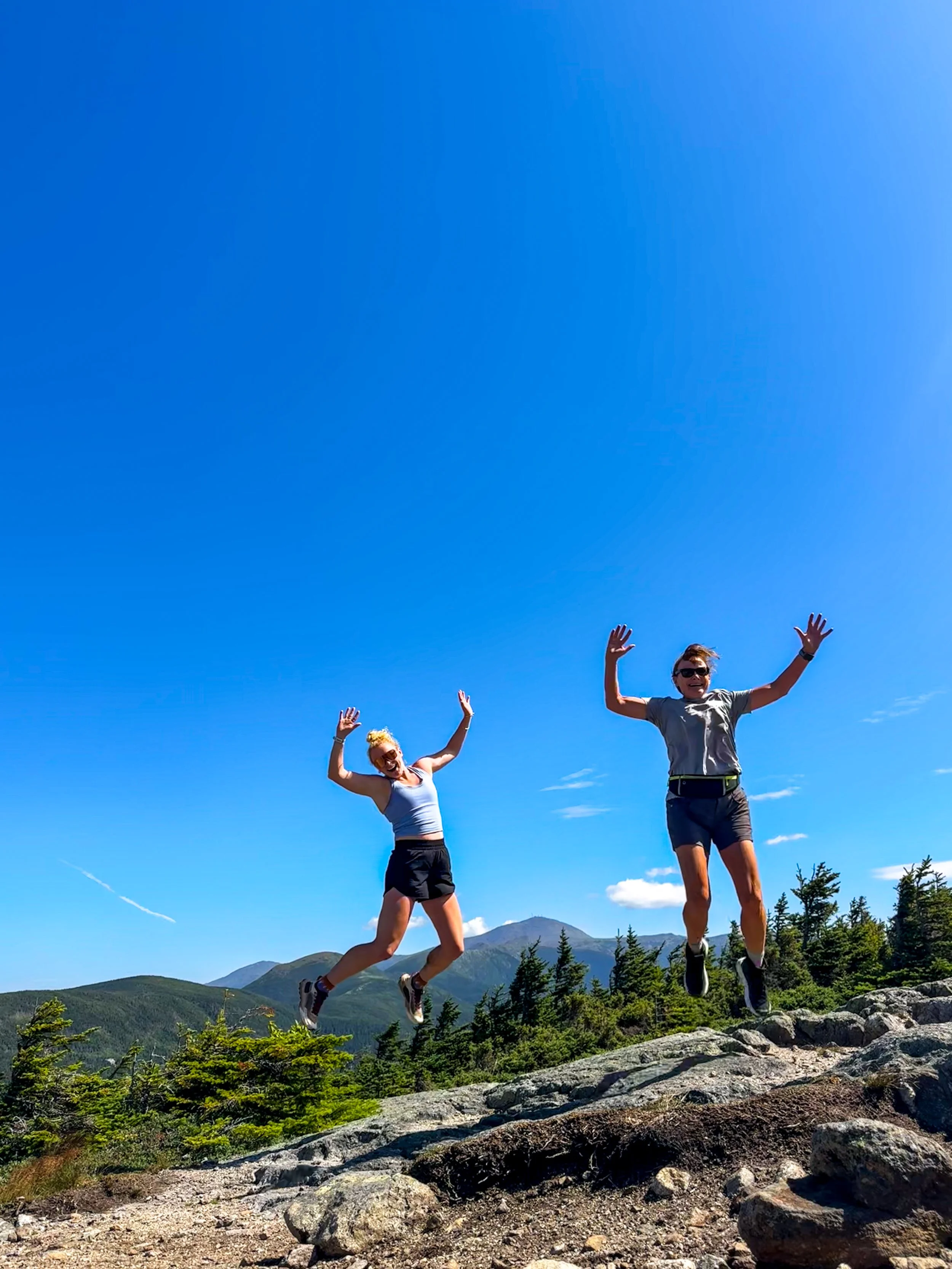 Two people jumping on a rocky trail in a mountainous landscape under a bright blue sky. Both are wearing athletic clothes and sunglasses, with arms raised in excitement.