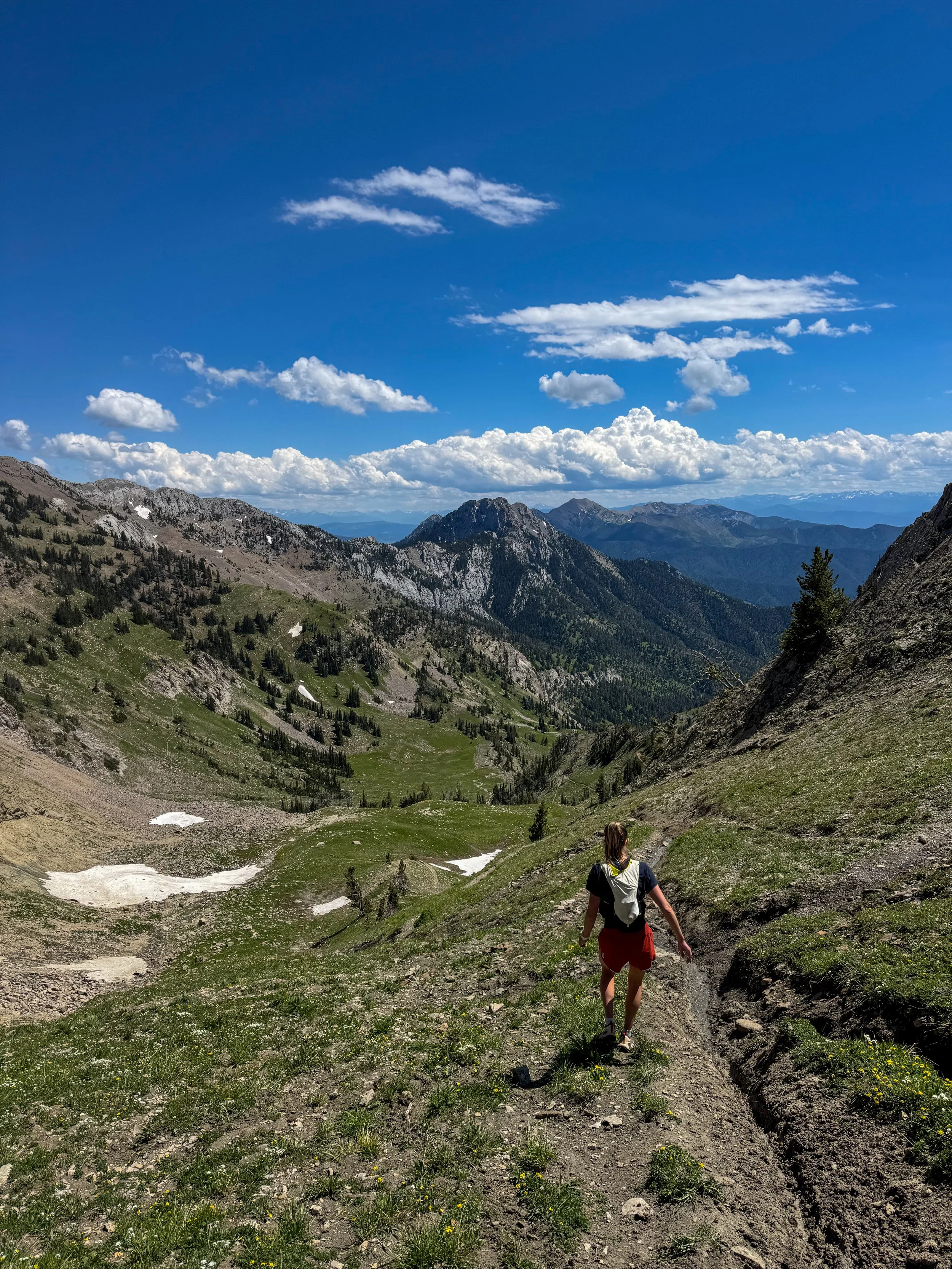 A person hiking on a mountain trail in a high-altitude area with green patches, snow remnants, and rugged mountain peaks under a partly cloudy blue sky.