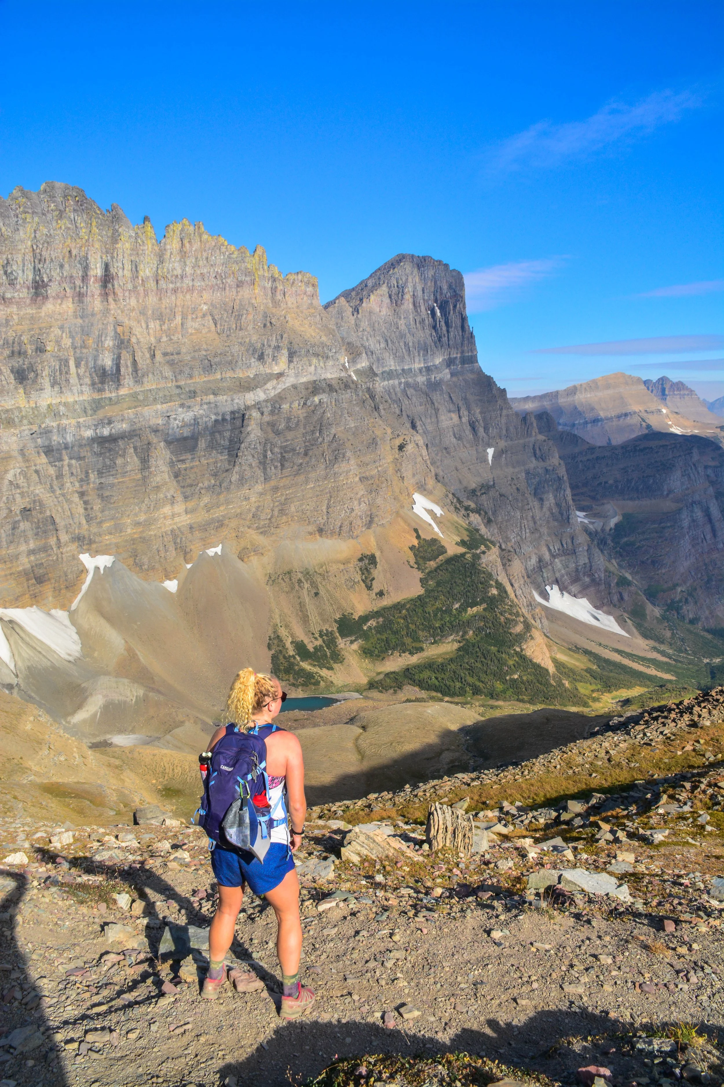 A woman with blonde hair, wearing a blue backpack, sunglasses, and hiking clothes, standing on a rocky trail and looking at a mountainous landscape with rugged cliffs, patches of snow, and a small lake in the distance under a bright blue sky.
