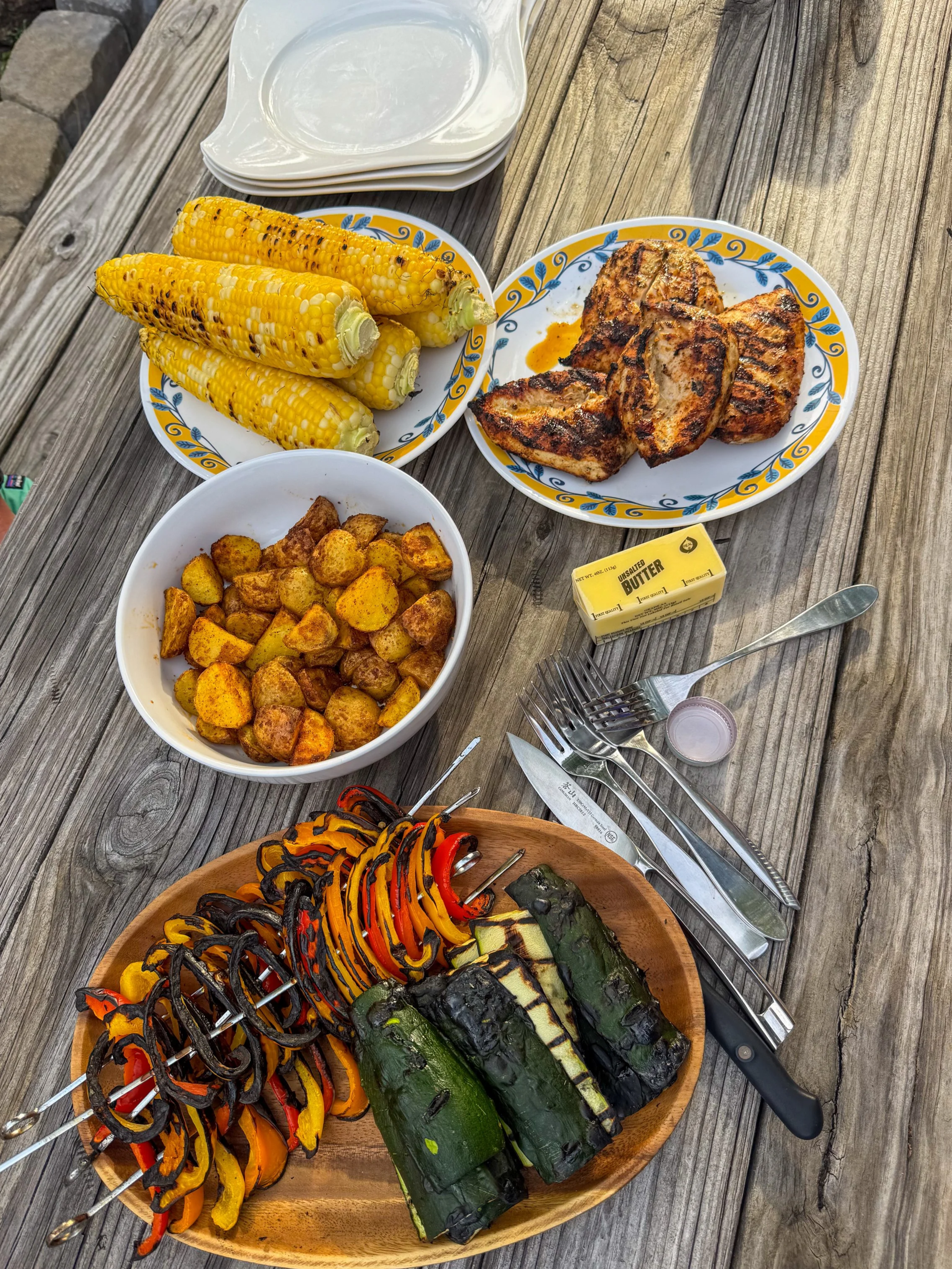 Grilled corn on the cob, roasted grilled pork chops, roasted potatoes, grilled vegetables skewers, and grilled zucchini and bell peppers on a wooden table.