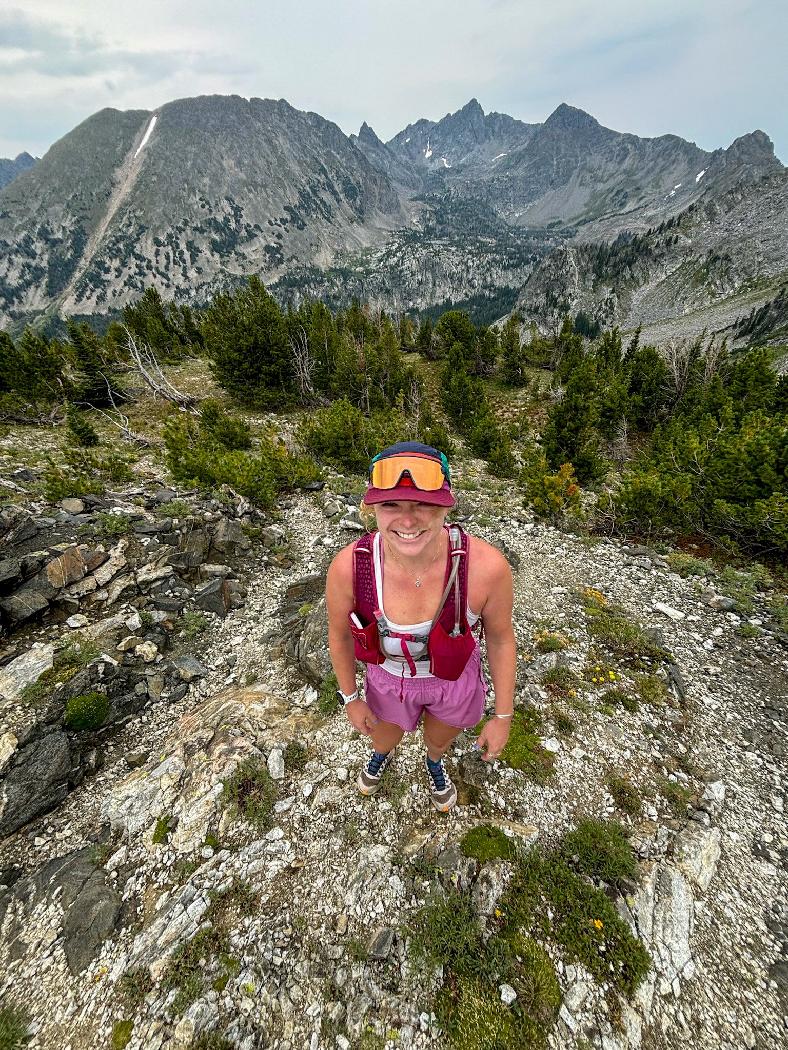 A smiling woman in hiking gear standing on a rocky trail in a mountainous area with green trees and rugged peaks in the background.