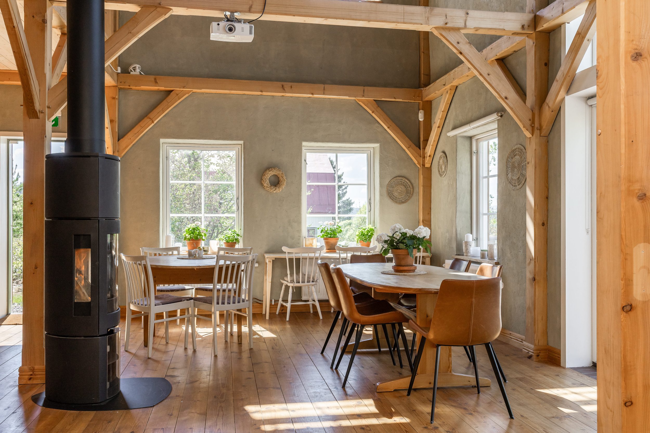 A cozy dining area with wooden beams, large windows, a wood stove, and a round table decorated with potted plants.