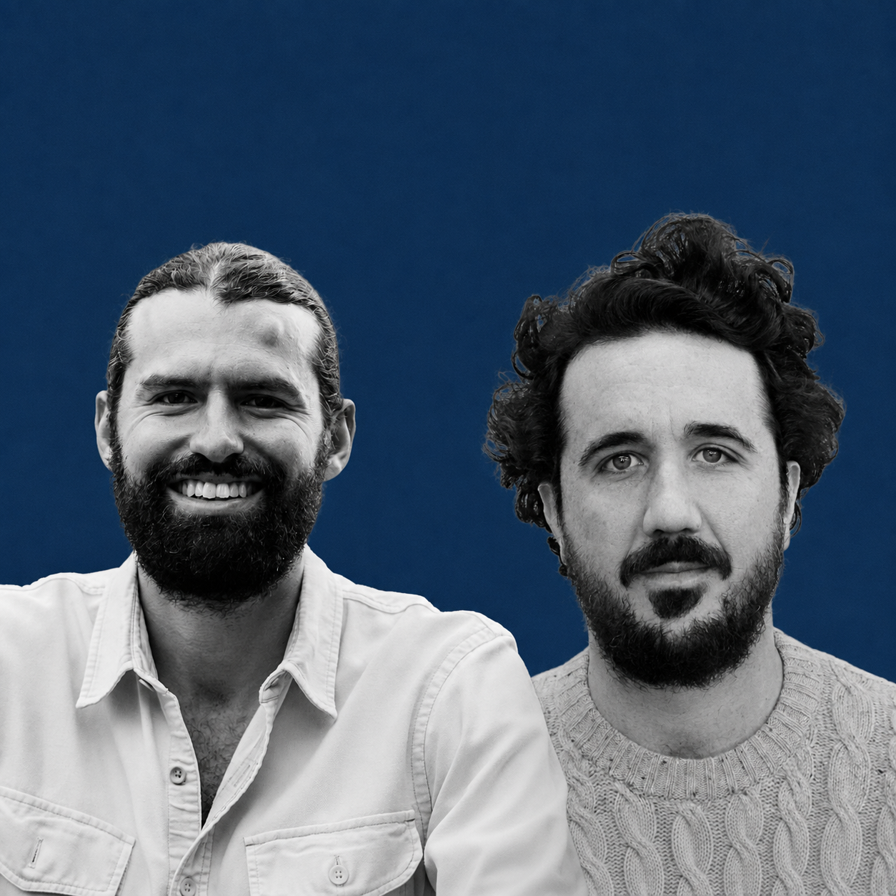Black and white portrait of two men, one with a beard and long hair, smiling, wearing a button-up shirt, and the other with curly hair and a beard, staring at the camera, against a dark solid background.