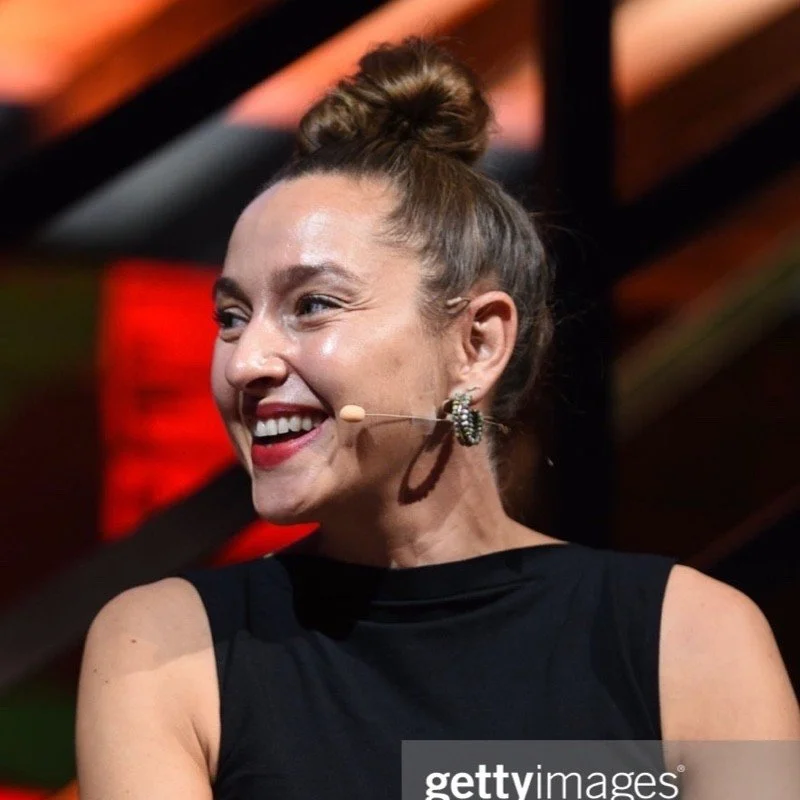 A woman with her hair in a bun, smiling and wearing a black sleeveless top, speaking into a headset microphone with a background of warm lighting and wooden beams.
