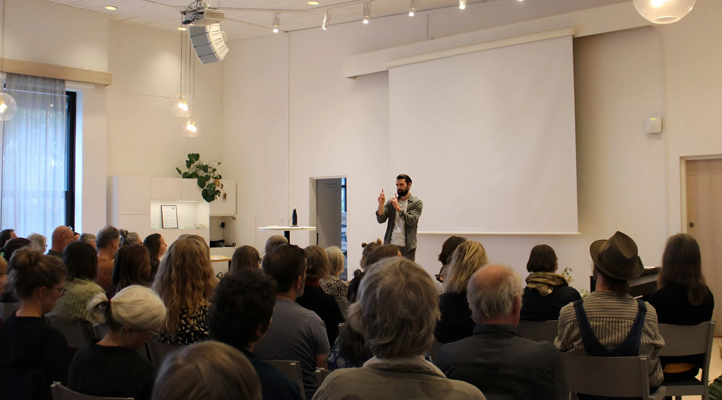 A man giving a presentation in front of a large audience in a bright, modern room with a white wall and ceiling. The audience is seated and attentively listening.