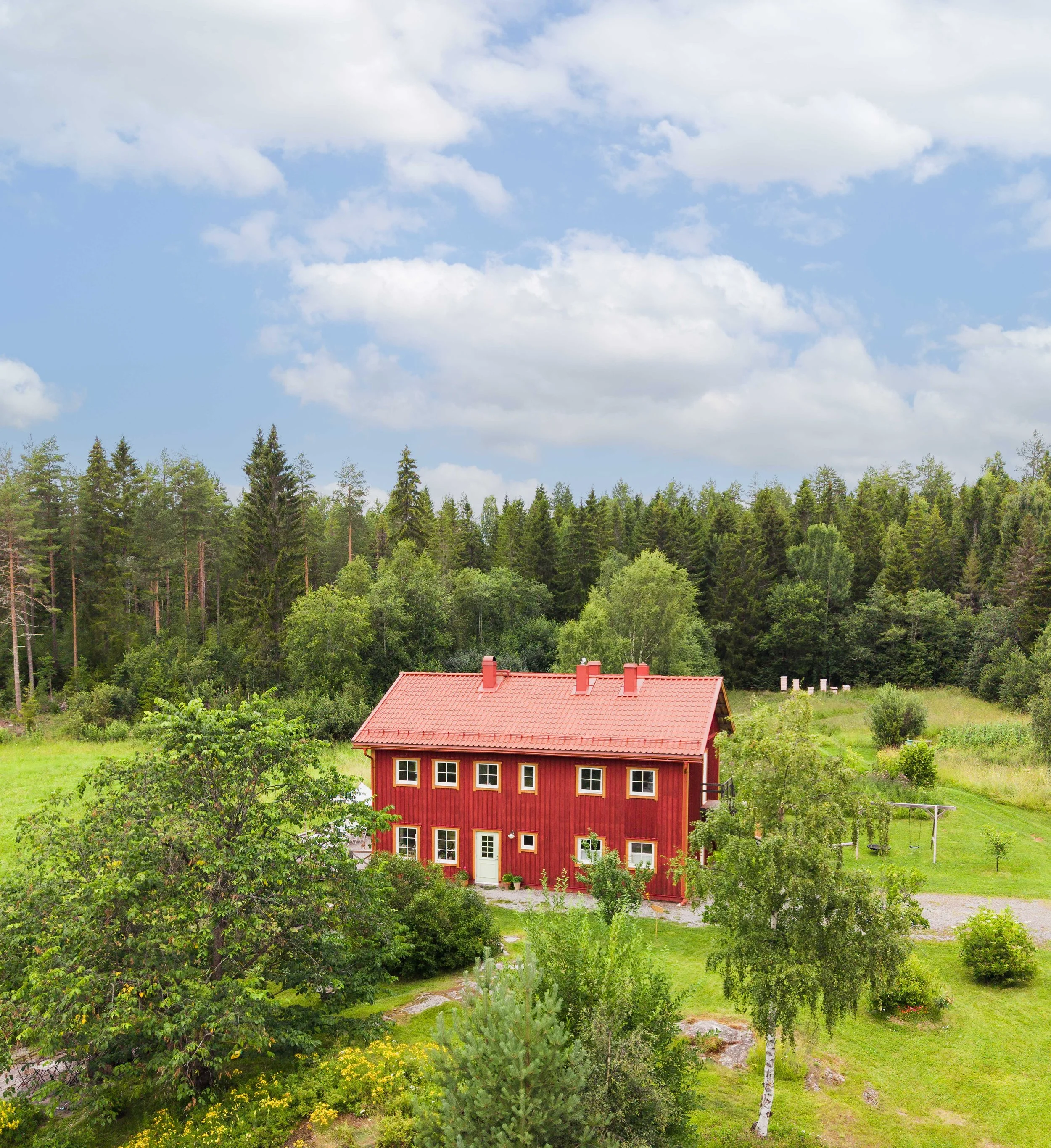 A red house with a red roof in a lush green landscape, surrounded by various trees and a grassy yard. A forest of tall trees is in the background, and the sky above is partly cloudy.