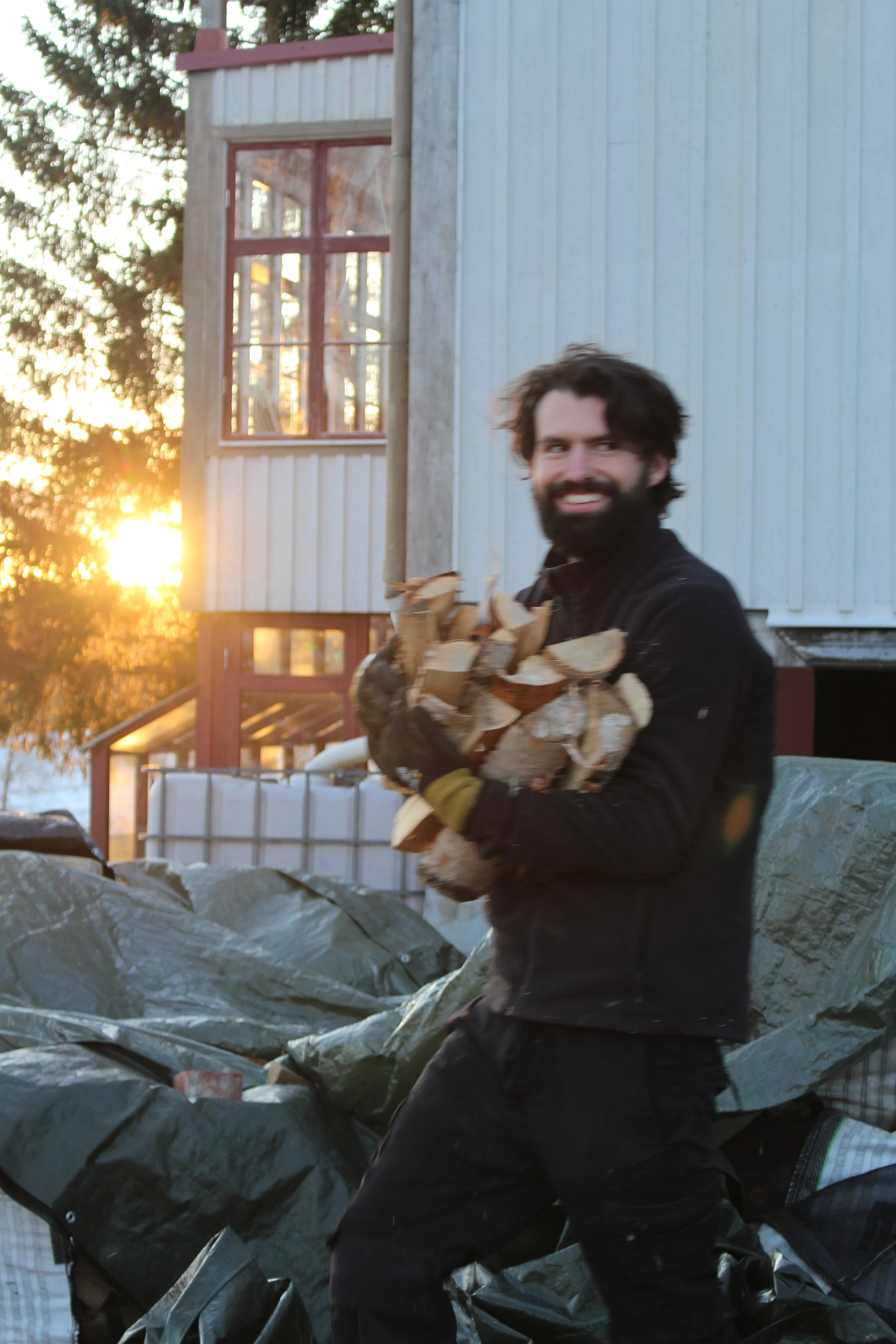 A man with a beard and dark curly hair holding a bundle of chopped wood outdoors during sunset, with a house and trees in the background.