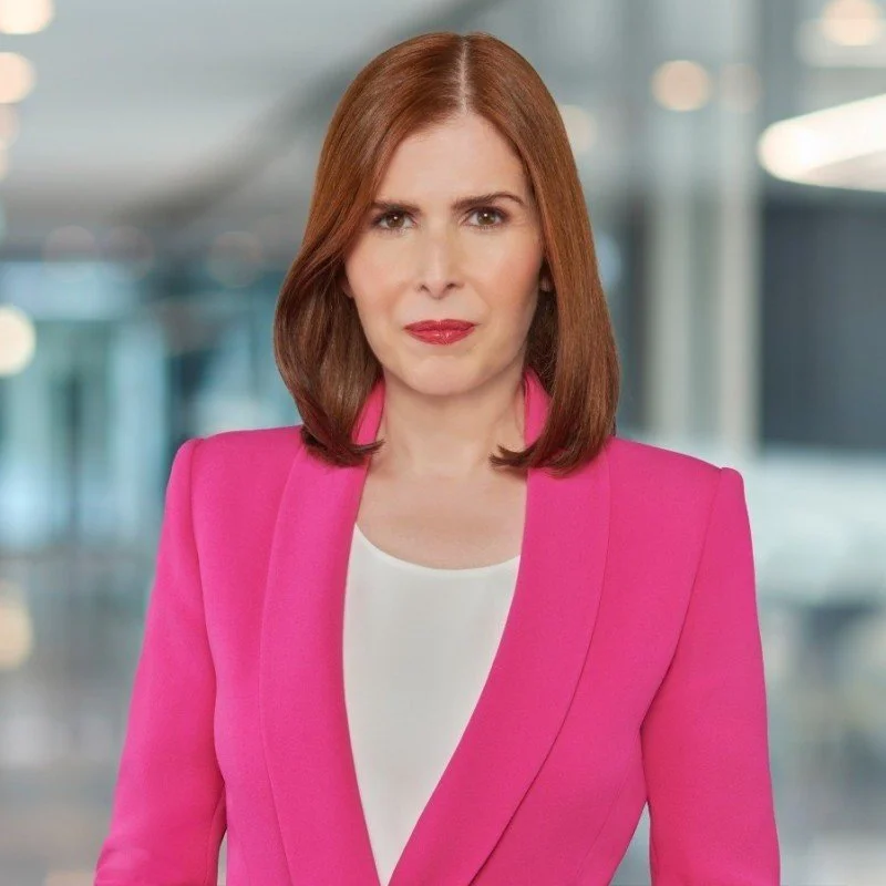 A woman with red hair wearing a pink blazer over a white top in a modern office environment.