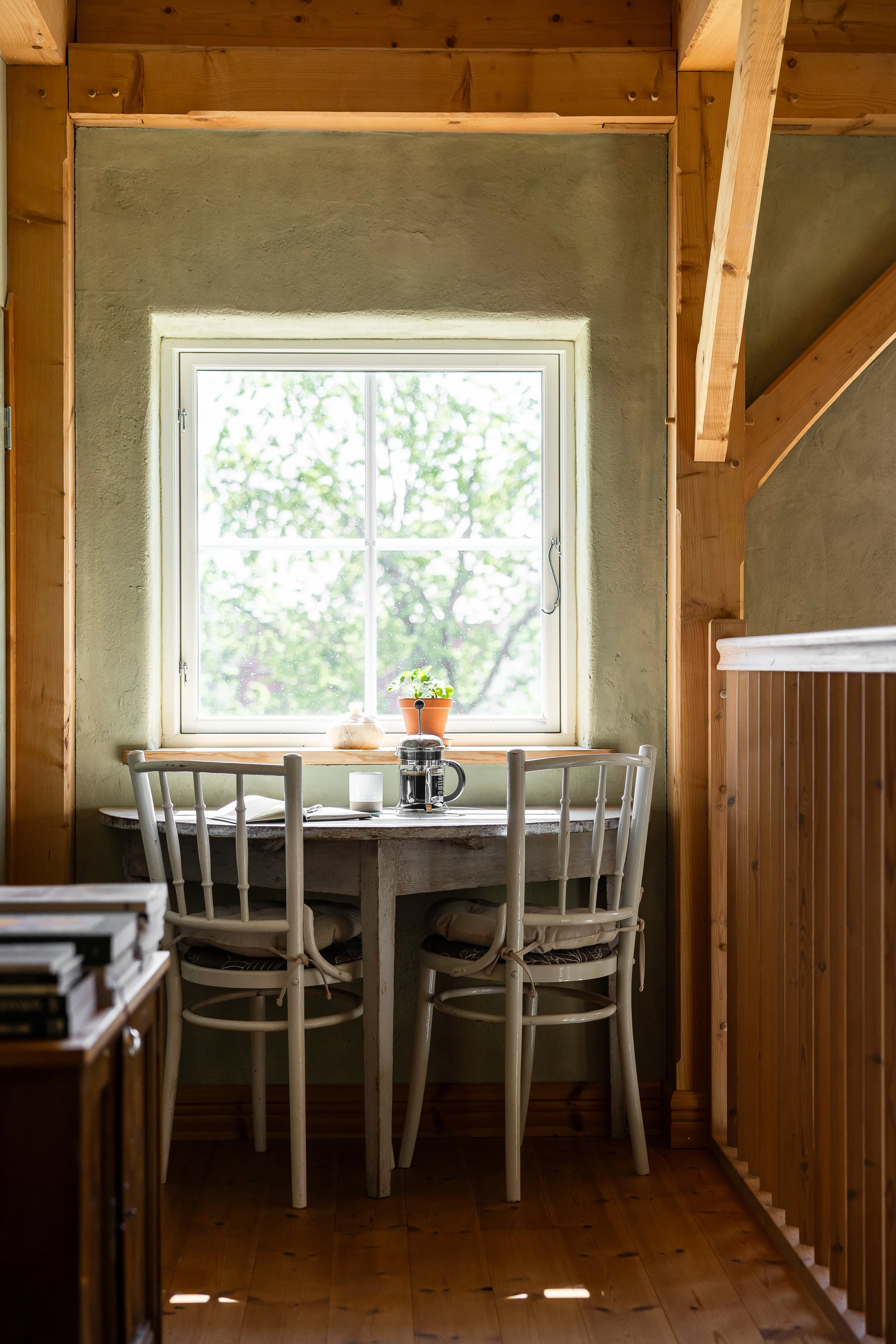 A cozy dining nook with a wooden table and six white chairs, set with a French press, cups, and a book, next to a large window with trees outside.