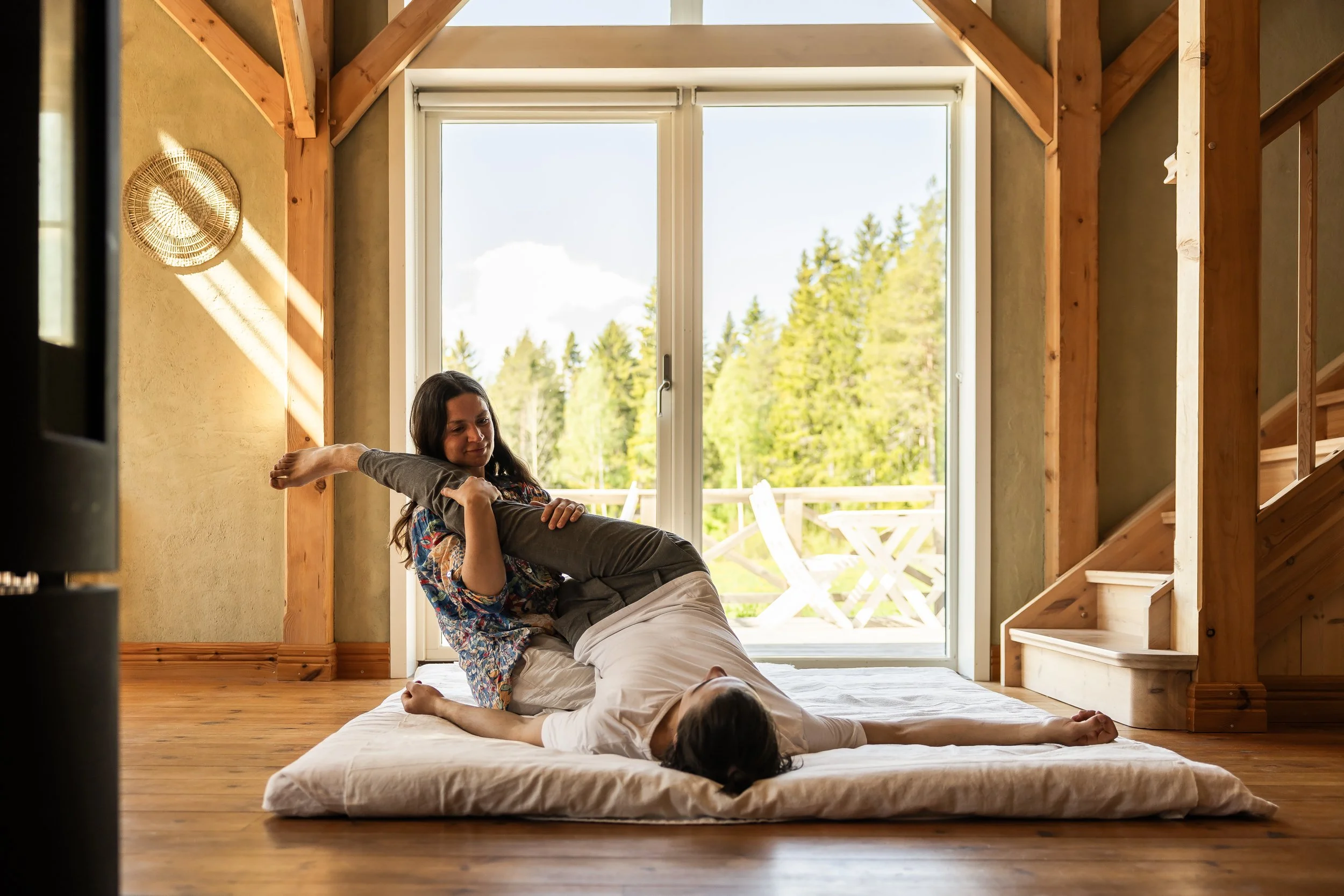 Two women practicing acro yoga on a mattress inside a wooden-framed room, with a large window showing trees outside.