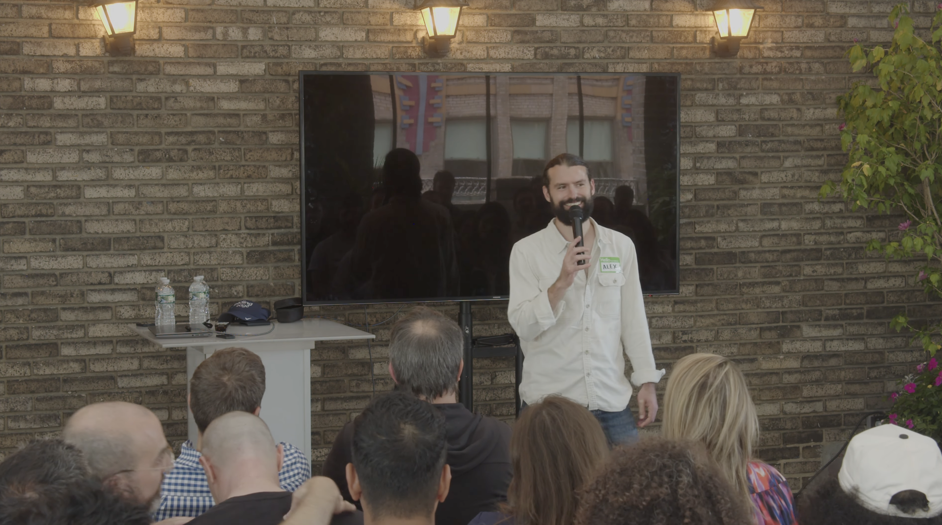 A man with a beard and long hair holding a microphone and speaking to a crowd during an event. He is wearing a white shirt with a name tag that says "Alex." Behind him, a large screen displays his reflection and the brick exterior of a building. The setting appears to be indoors with an audience seated in front.
