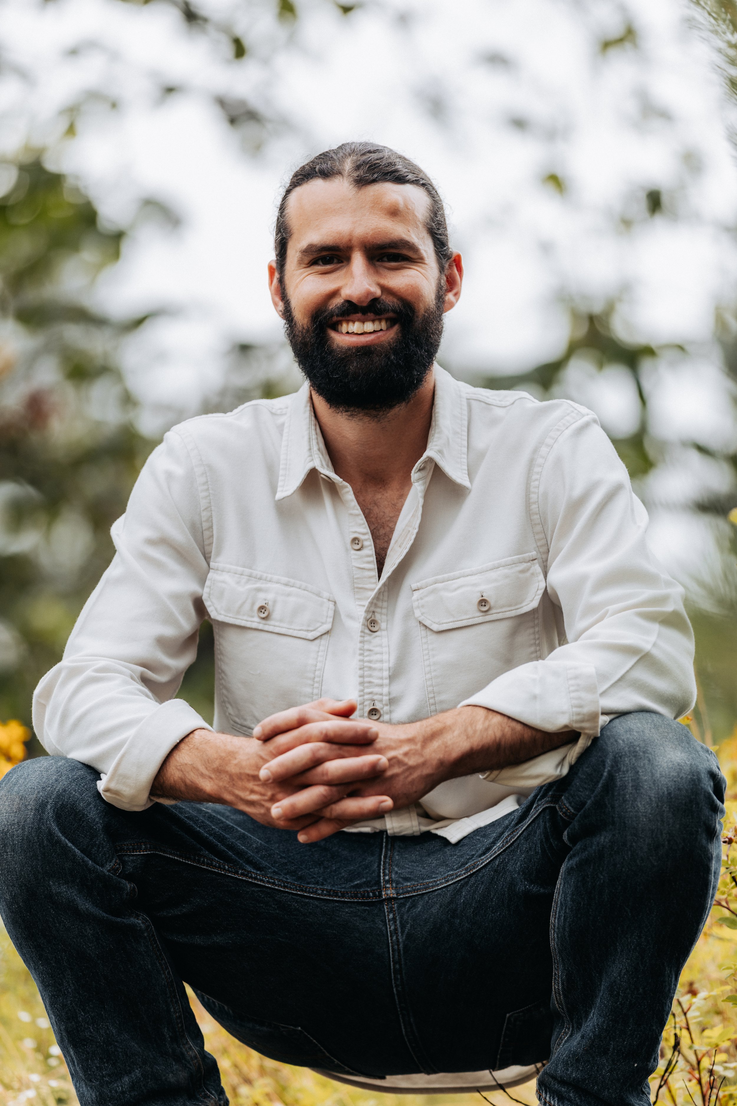A man with a beard and long hair sitting outdoors in a park or garden, smiling at the camera.