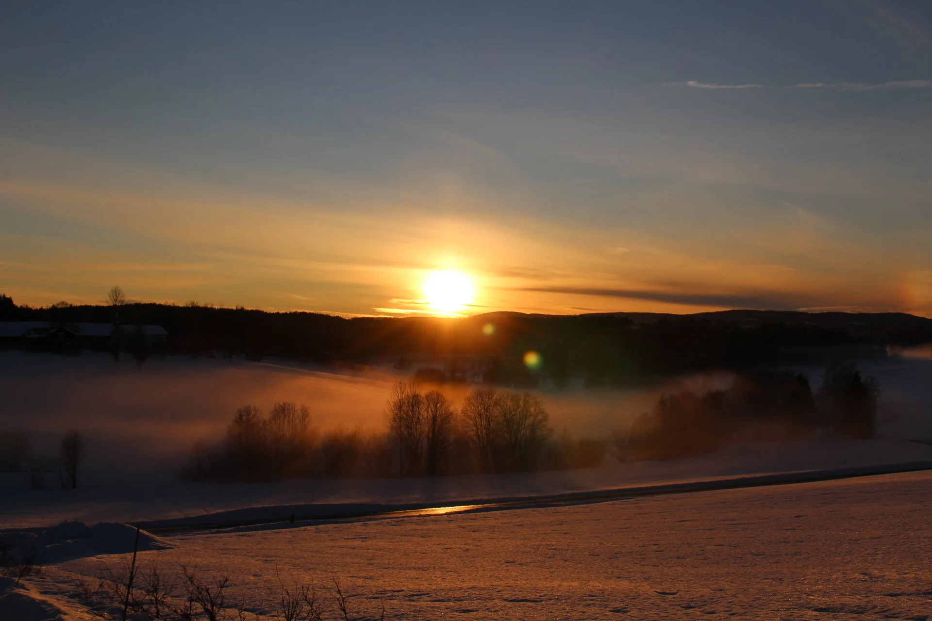 Sunset over a snow-covered landscape with fog, trees, and rolling hills in the distance.