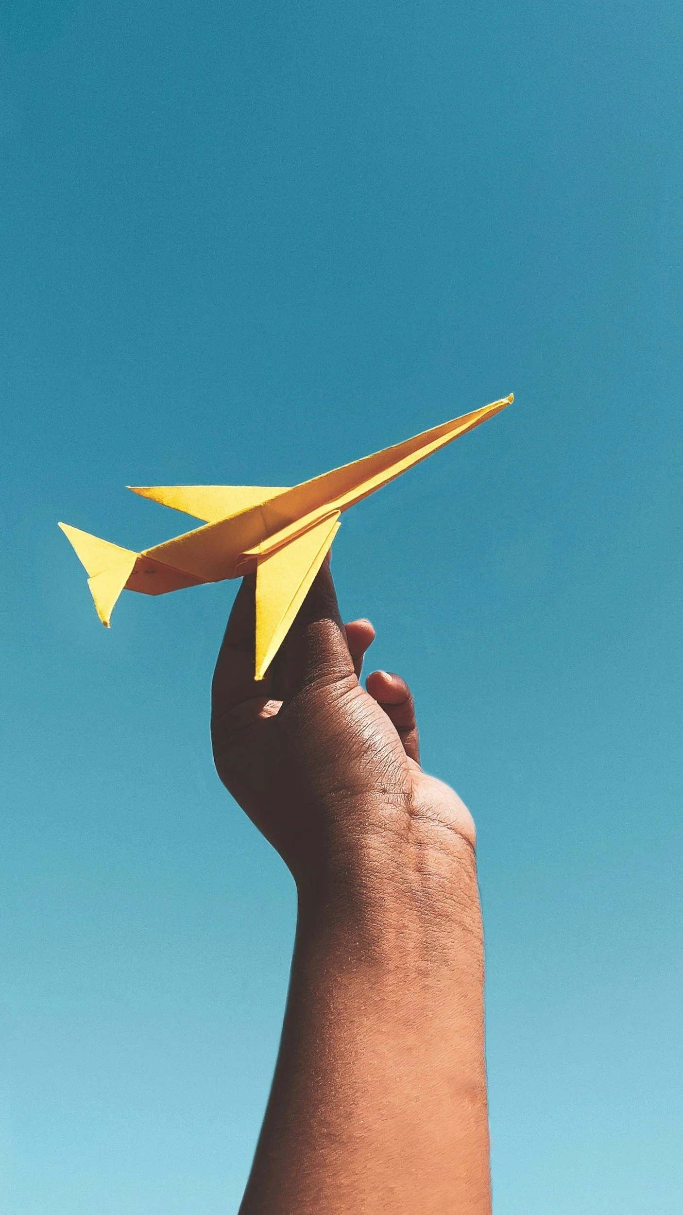 A person holding a yellow paper airplane against a clear blue sky.