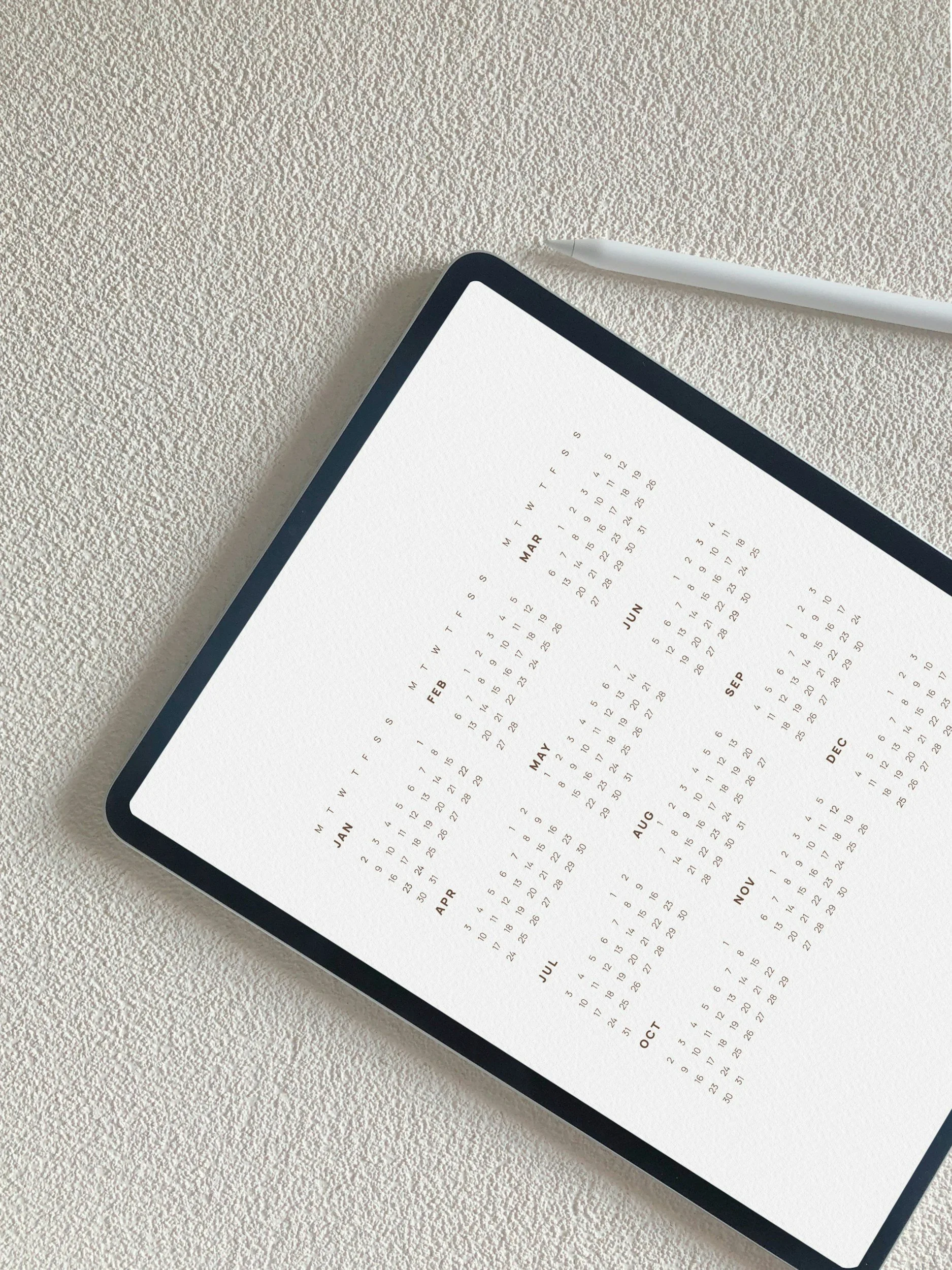 A digital tablet displaying a yearly calendar with months from January to December on a textured white surface, with a white stylus pen nearby.
