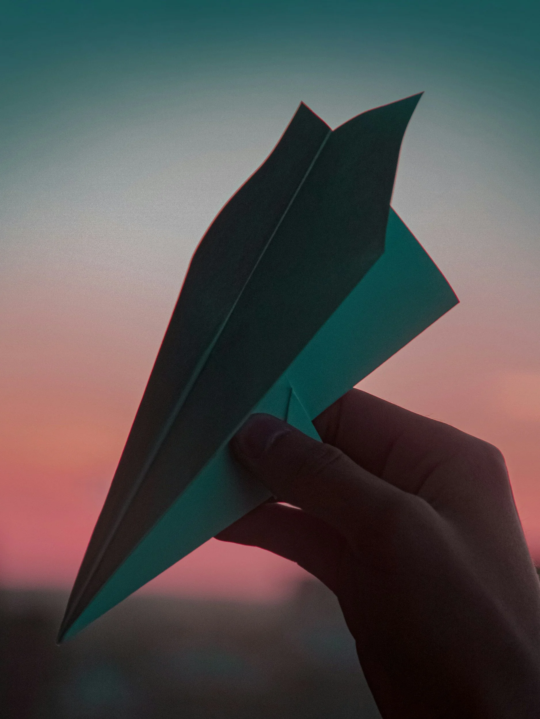 A hand holding an open book or notebook against a colorful, gradient sky during sunset or twilight.