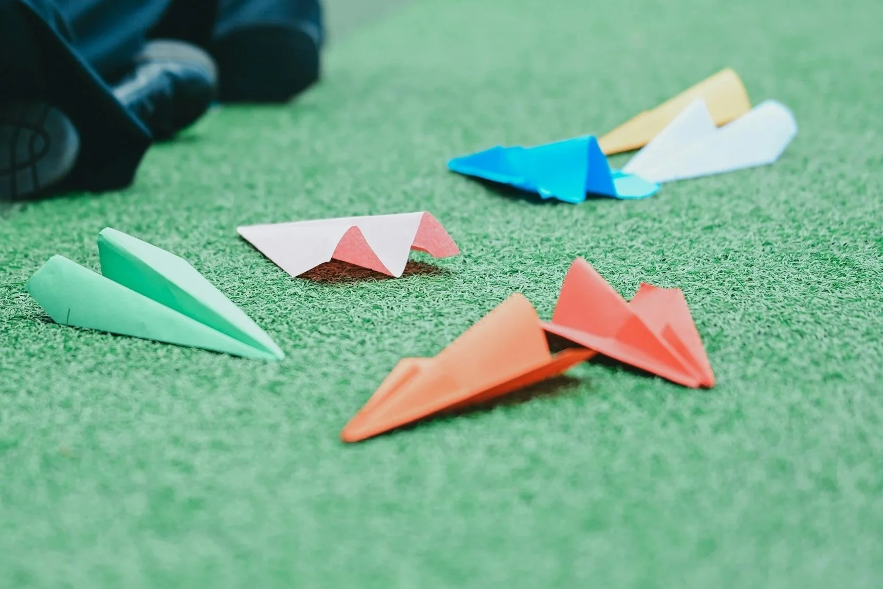 Colorful origami paper airplanes on green carpet, with a person's shoe visible in the background.