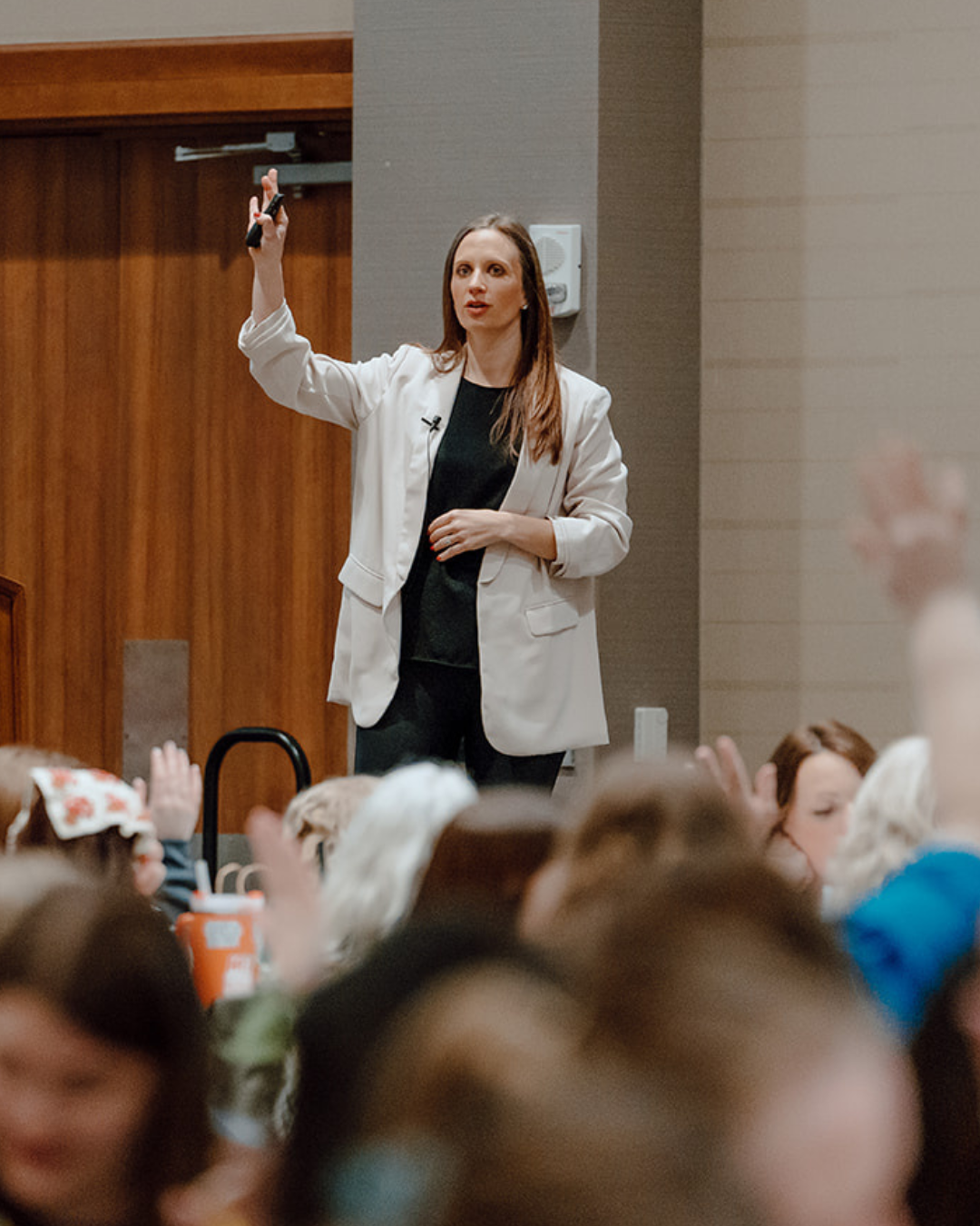 A woman giving a presentation at a conference to an audience, gesturing with her right hand, with multiple people watching.