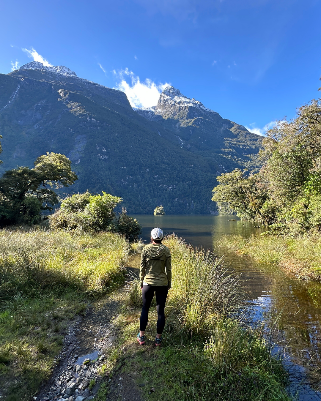 Person walking along a narrow trail beside a lake with lush green trees, tall mountains with snow-capped peaks in the background, and a clear blue sky.