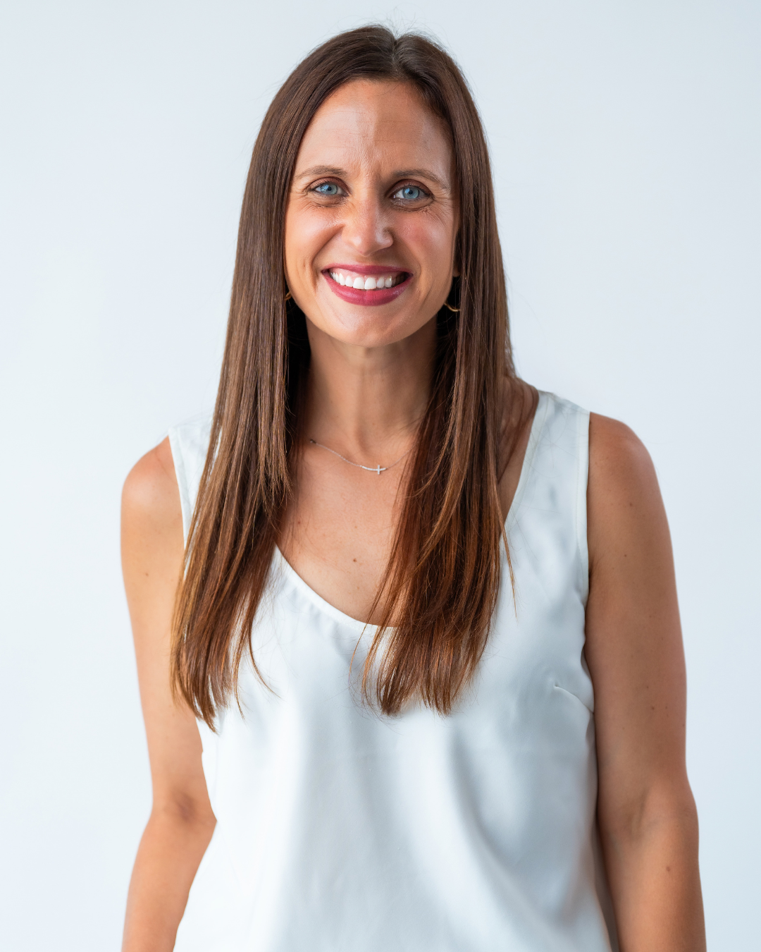 A woman with long brown hair, blue eyes, and a bright smile, wearing a white sleeveless top and a delicate cross necklace, standing against a plain light background.
