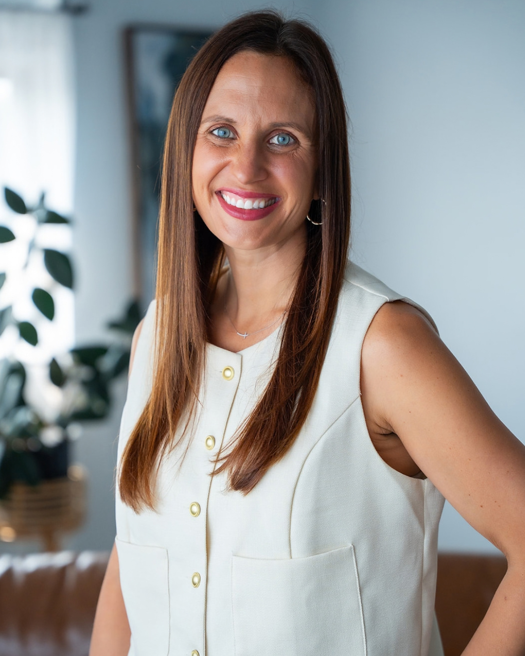A woman with long brown hair and blue eyes, smiling and wearing a sleeveless cream-colored top with gold buttons, standing indoors with a plant and picture frame in the background.