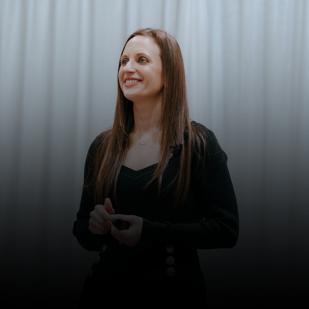 A woman with long brown hair smiling during a presentation, holding a remote control in her hands, standing against a plain, light-colored curtain background.