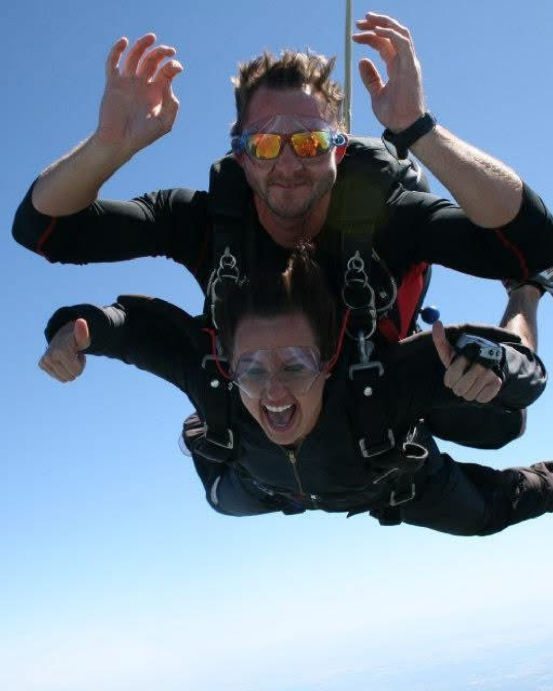 Two people skydiving, one man with sunglasses and a woman with goggles, smiling and enjoying the free fall against a clear blue sky.