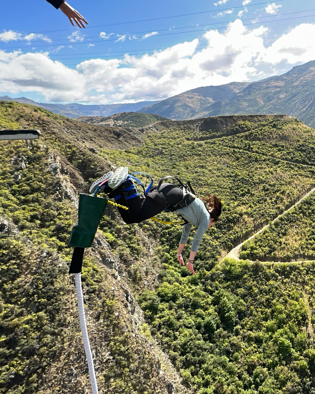 A person appears to be falling or hanging off a cliff, with a harness and equipment on, overlooking a mountainous landscape with green hills and a partly cloudy sky.