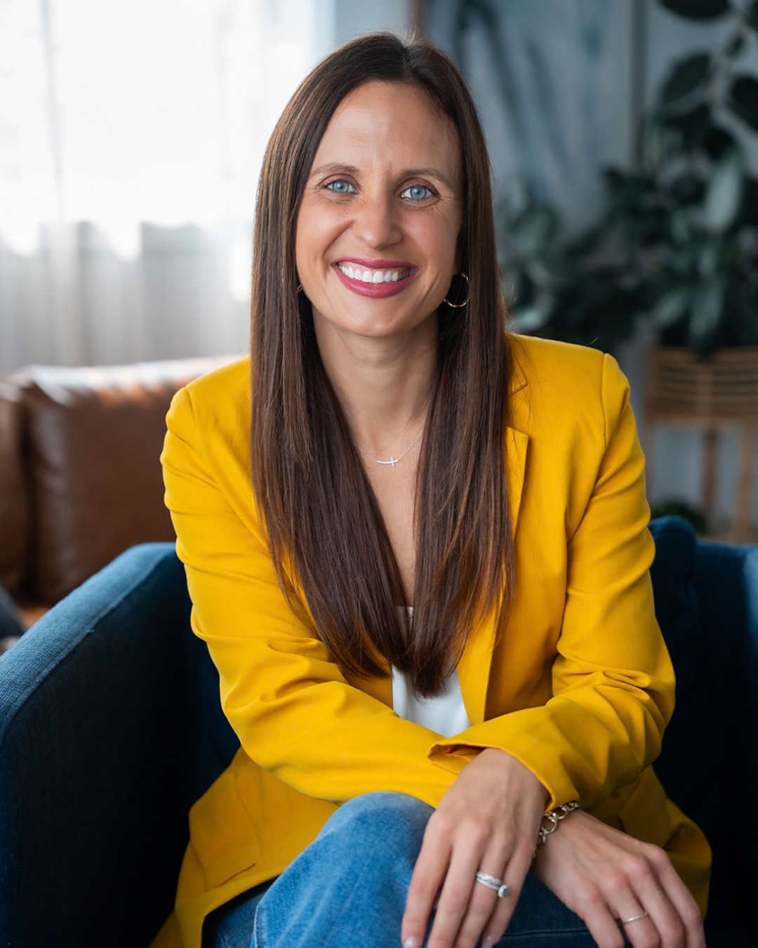 A woman with long brown hair, blue eyes, and light skin, smiling, wearing a yellow blazer and white top, seated indoors in a living room with a couch and plants in the background.