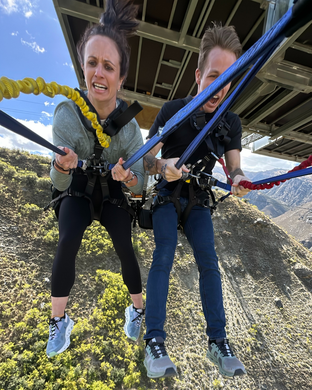 Two people on a zip line in mid-air, holding onto the safety handles with mountains and greenery in the background, and a partly cloudy sky above.