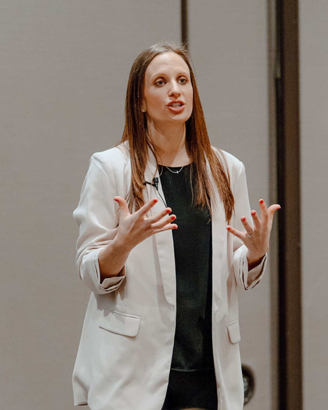 A woman with long reddish-brown hair, wearing a white blazer over a black top, speaking and gesturing with her hands during a presentation or speech.