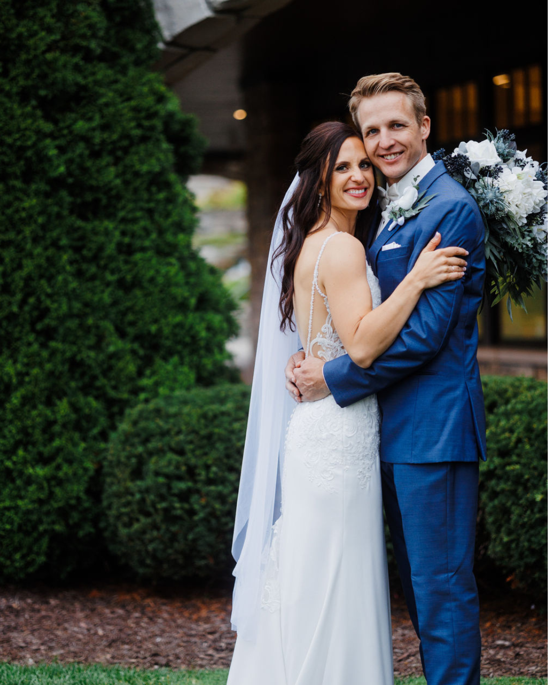 A bride and groom smiling and hugging outdoors, bride in a white wedding dress with lace details, groom in a blue suit, with greenery and a building in the background.
