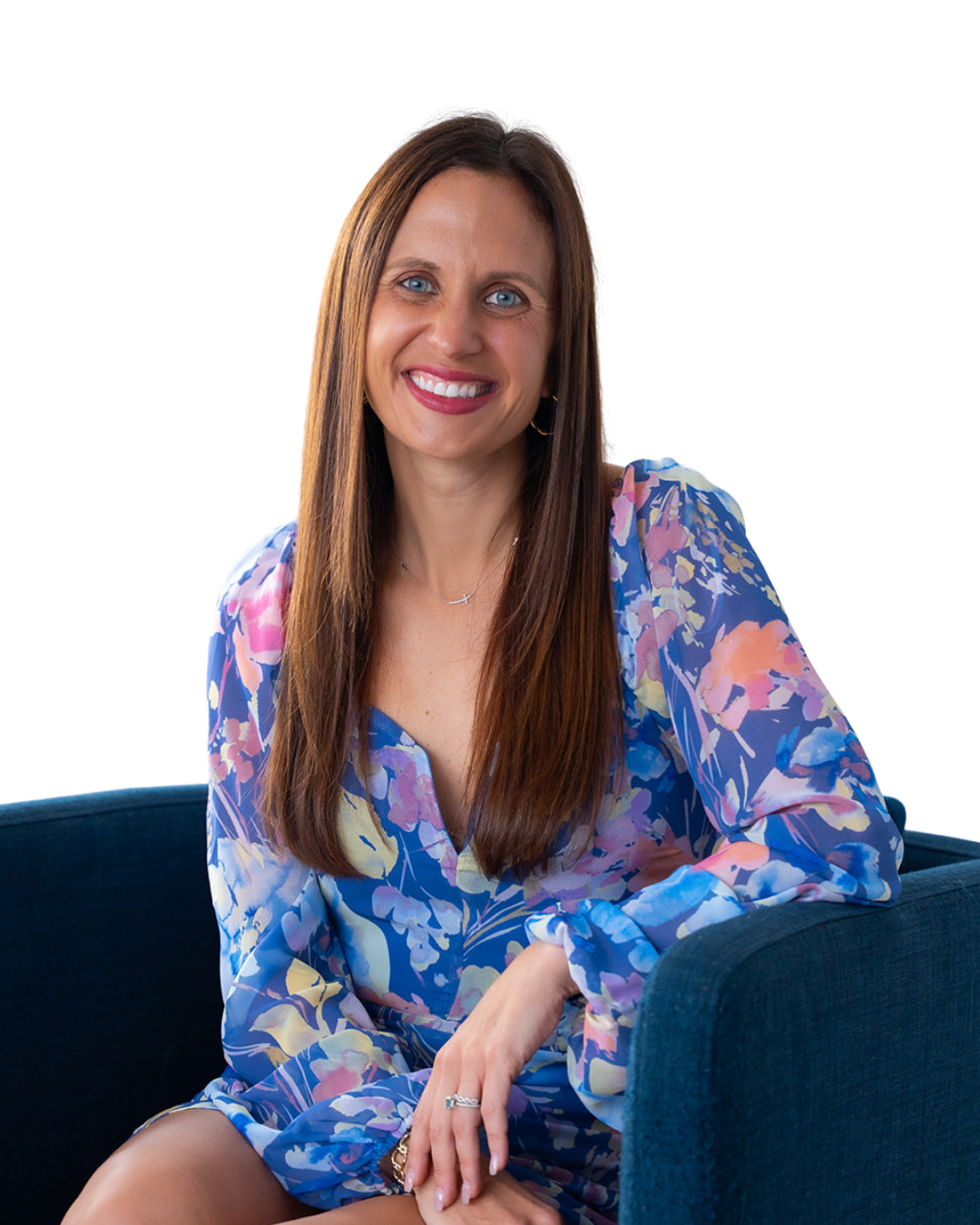 A woman with long brown hair and blue eyes, smiling, sitting on a blue couch, wearing a colorful floral dress.