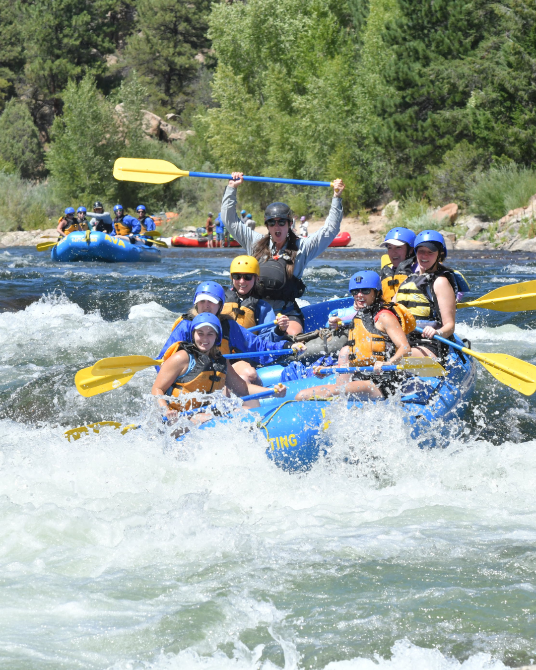 Group of people white water rafting on a river, wearing life jackets and helmets, with one woman excitedly holding a paddle above her head.