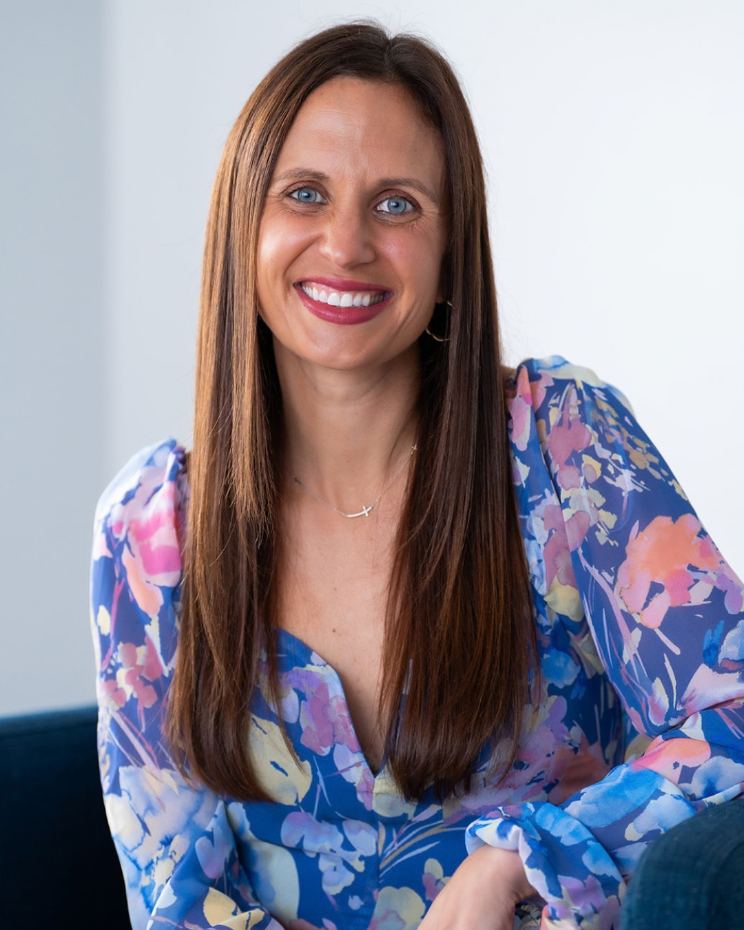 A woman with long brown hair, blue eyes, and a bright smile, wearing a colorful floral blouse, sitting on a dark-colored piece of furniture with a white background.